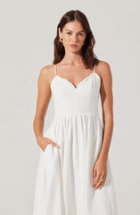 A woman is wearing a white sleeveless dress with thin spaghetti straps. The dress features a fitted bodice with a sweetheart neckline and a gathered, flowing skirt. She has her right hand in her pocket and is accessorized with large, gold hoop earrings and a gold ring on her left hand.