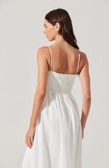A woman is wearing a sleeveless white dress with thin spaghetti straps. The dress has a smocked bodice and a flowing skirt. She has long, dark, wavy hair and is wearing gold hoop earrings.