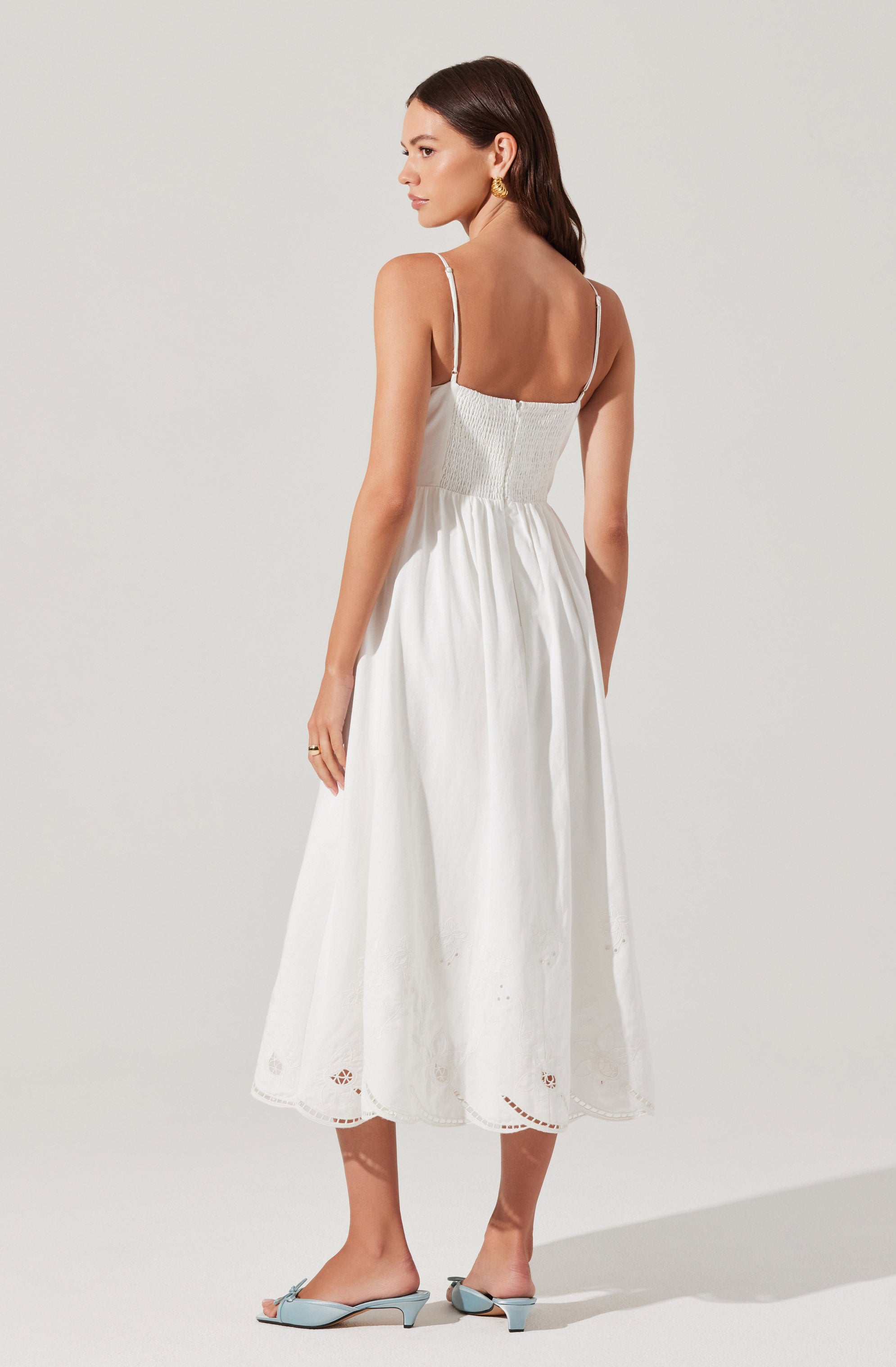 A woman is wearing a white, sleeveless, ankle-length dress with thin shoulder straps and embroidered detailing at the hem. She is also wearing matching light blue open-toe mules.