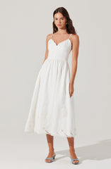 A woman is wearing a white, sleeveless, fit-and-flare maxi dress with a sweetheart neckline and thin spaghetti straps. The dress features gathered detail at the waist and has delicate eyelet embroidery along the hem. She pairs the outfit with open-toe heels and gold earrings.