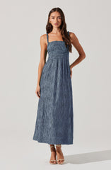A woman is wearing a blue sleeveless maxi dress with adjustable straps. The dress features a smocked bodice with a fitted waist and a long, flowing skirt made of textured, crinkled fabric. She is also wearing tan high-heeled sandals.