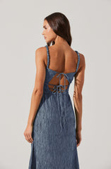 A woman is wearing a sleeveless blue dress with textured fabric. The dress features shoulder straps with ruffled edges, an open back with crisscrossing adjustable ties, and a flowing skirt that reaches the floor.