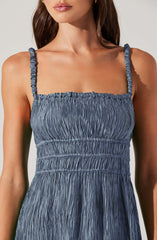 A woman is wearing a blue sleeveless dress with a textured, crinkled fabric. The dress has thin, gathered straps and a fitted bodice with horizontal ruching, flowing into a gathered skirt.