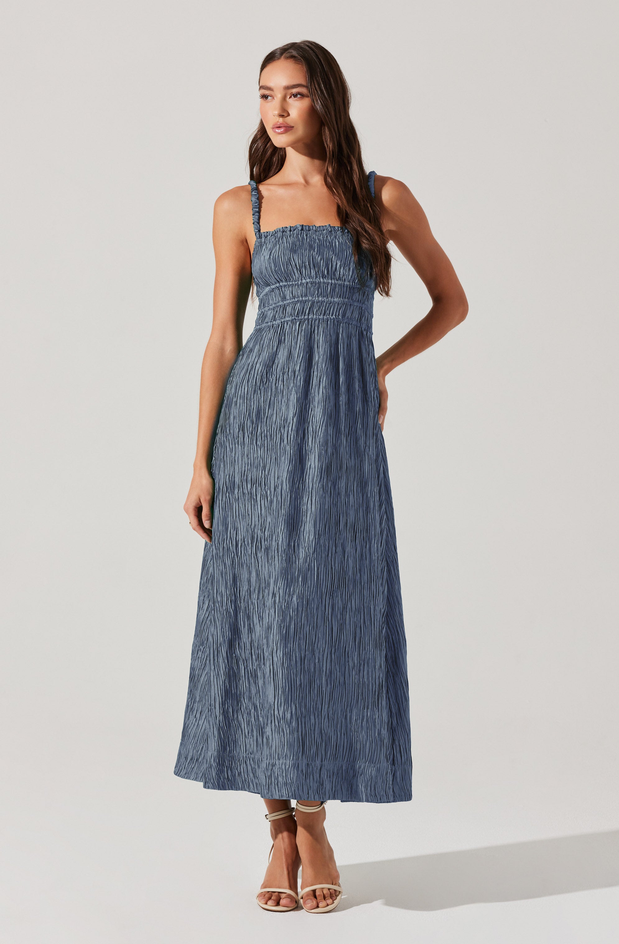 A woman is wearing a sleeveless, full-length, blue crinkled maxi dress with thin straps. The dress has a fitted bodice with shirred detailing and a flowing skirt. She is also wearing strappy beige high-heeled sandals.