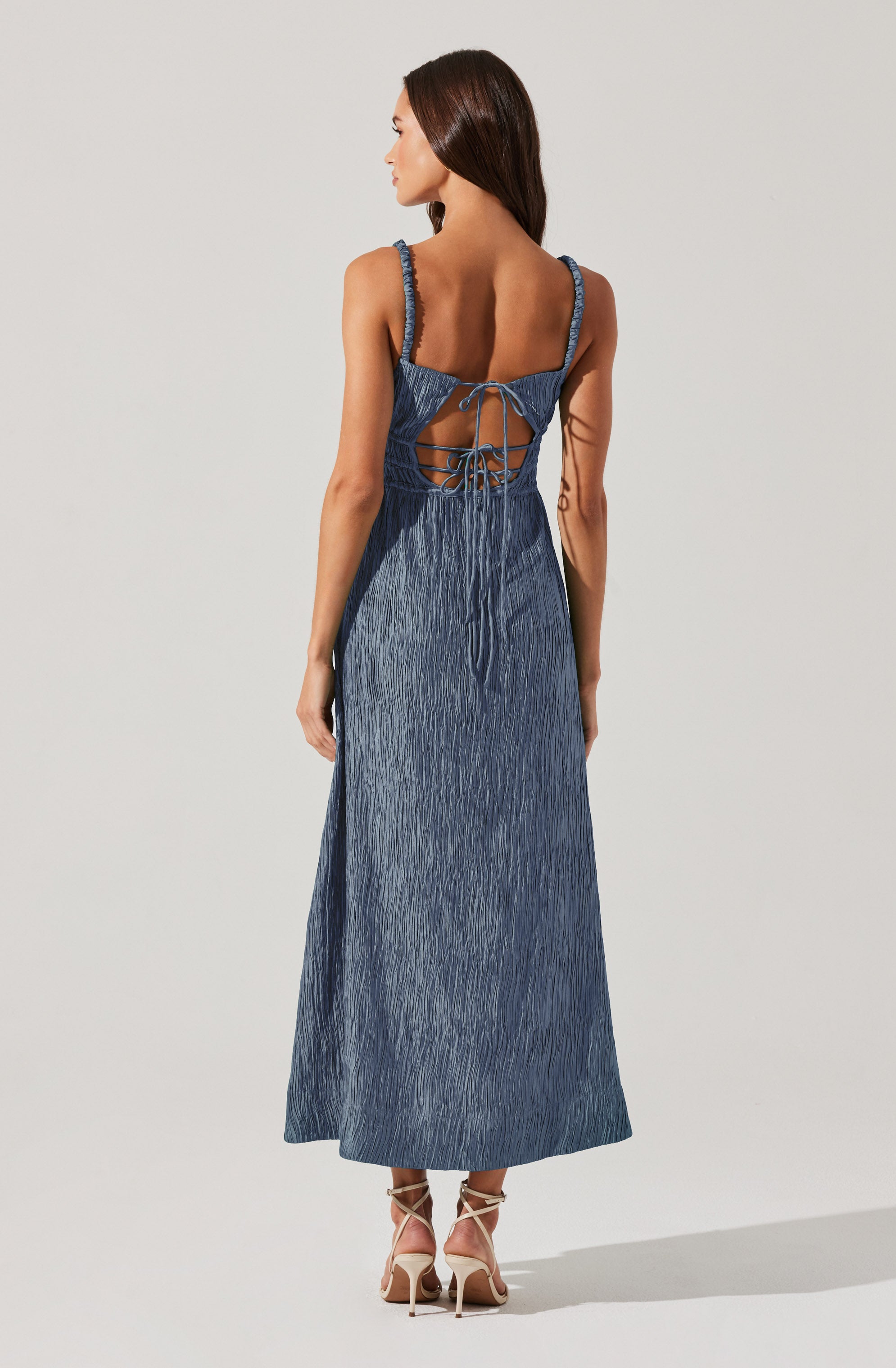 A woman is wearing a long, sleeveless denim dress with textured fabric. The dress features braided shoulder straps and an open back with crisscross laces tied in a bow. The hem reaches down to her ankles, and she is wearing high-heeled sandals with ankle straps.