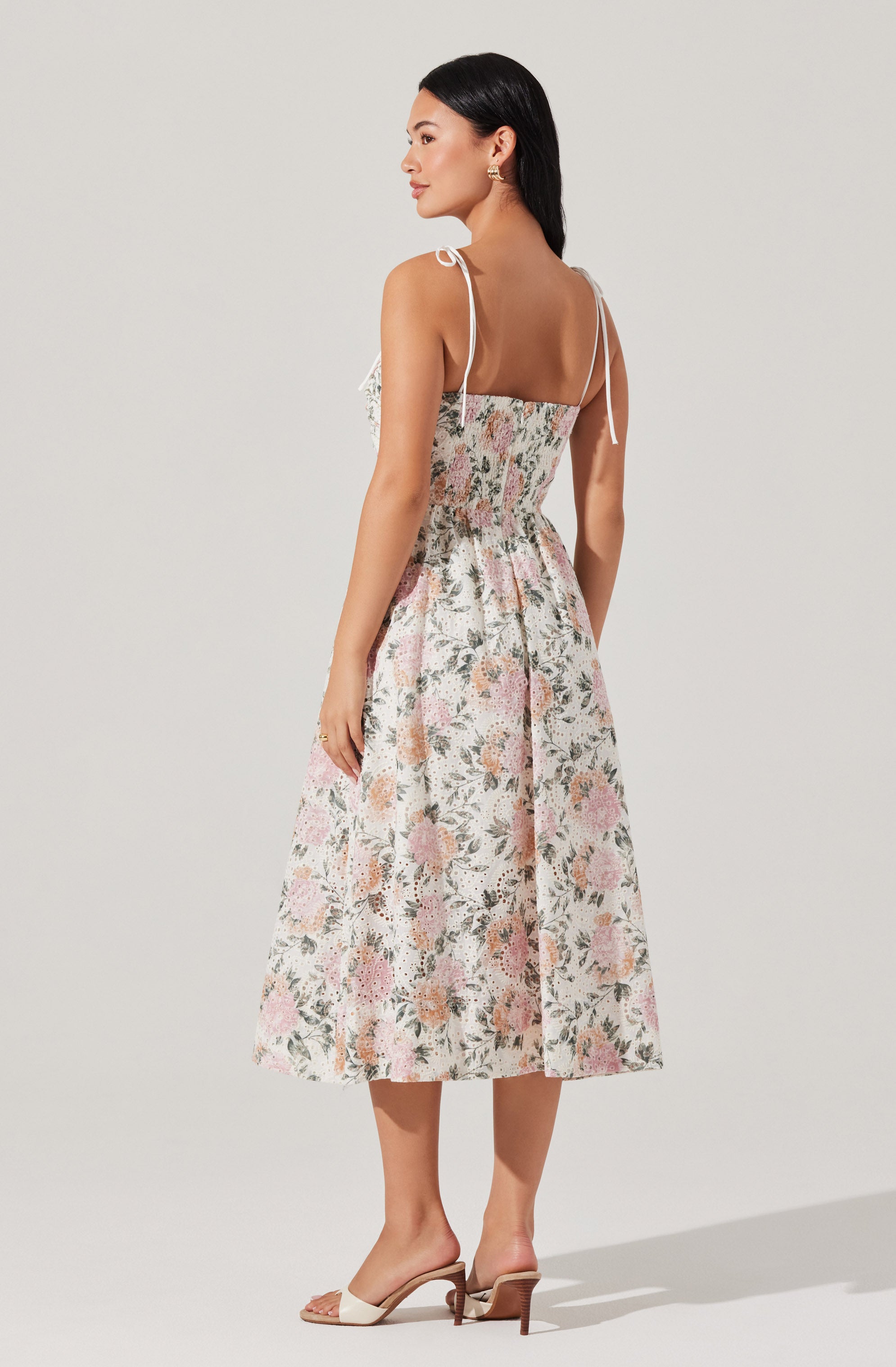 The dress is a sleeveless, midi-length style with thin, adjustable shoulder straps tied into bows. It features a fitted bodice with a smocked detail at the waist that extends to the skirt, which has a gentle A-line shape. The fabric has a floral pattern with soft pink, green, and beige tones on a light background. The dress has a modest neckline and a relaxed, flowy silhouette. The model is wearing beige open-toe heeled sandals.