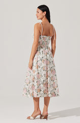 The dress is a sleeveless, midi-length style with thin, adjustable shoulder straps tied into bows. It features a fitted bodice with a smocked detail at the waist that extends to the skirt, which has a gentle A-line shape. The fabric has a floral pattern with soft pink, green, and beige tones on a light background. The dress has a modest neckline and a relaxed, flowy silhouette. The model is wearing beige open-toe heeled sandals.