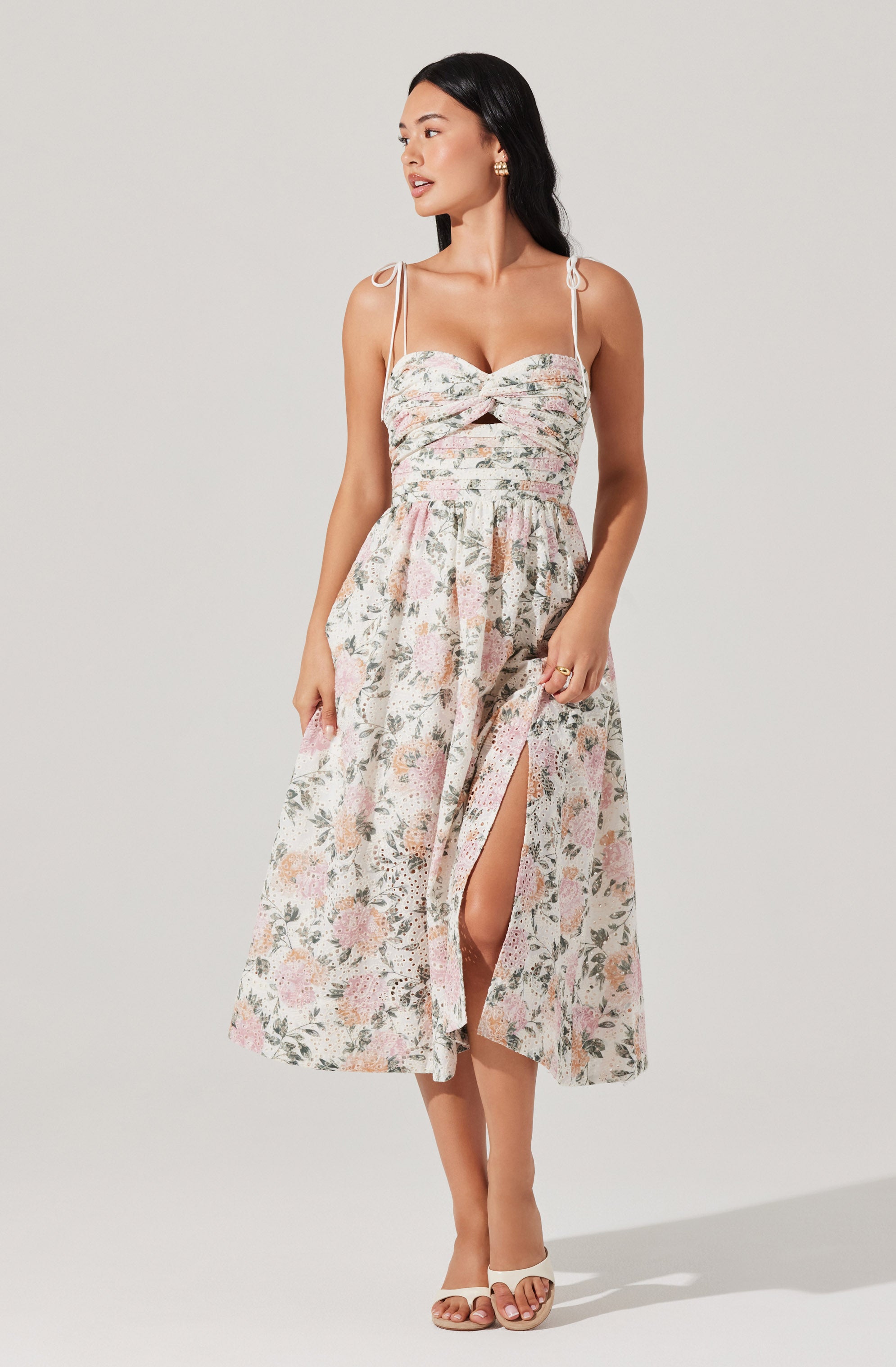 A woman is wearing a sleeveless dress with thin off-shoulder straps tied into bows on her shoulders. The dress has a fitted bodice with a twisted knot detail at the bust and a cutout below it. The skirt is long, flowy, and features a floral print with soft pastel colors. There is a slit on the front of the skirt revealing part of her leg. She is wearing open-toe sandals and has her dark hair down.