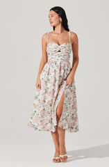 A woman is wearing a sleeveless dress with thin off-shoulder straps tied into bows on her shoulders. The dress has a fitted bodice with a twisted knot detail at the bust and a cutout below it. The skirt is long, flowy, and features a floral print with soft pastel colors. There is a slit on the front of the skirt revealing part of her leg. She is wearing open-toe sandals and has her dark hair down.