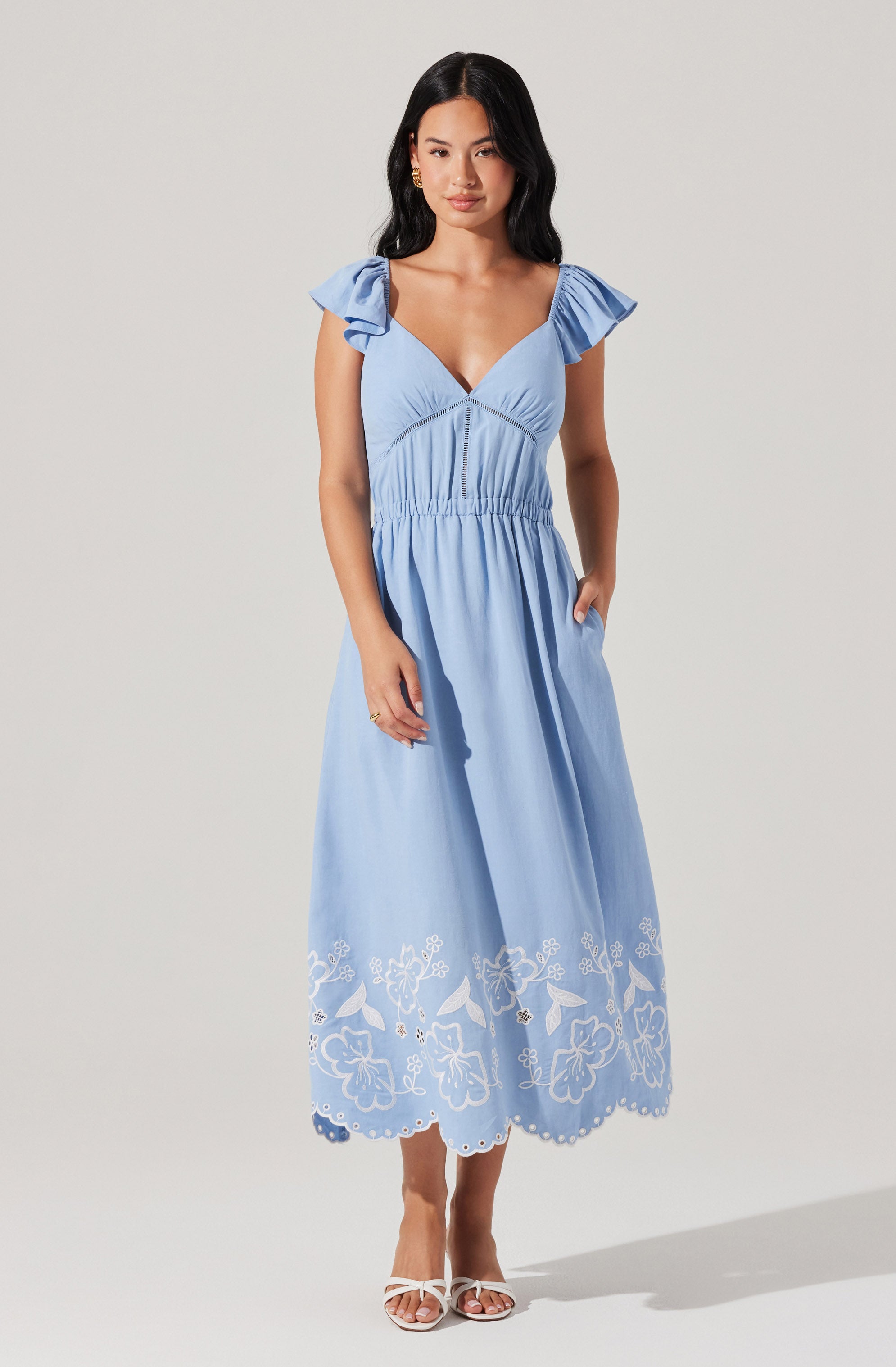 A woman is wearing a light blue, sleeveless, V-neck maxi dress with ruffled shoulder details. The dress has a gathered waistline and features an embroidered floral and scalloped hem pattern in white at the bottom. She is also wearing white open-toed heels.