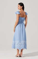 A woman is wearing a light blue, midi-length dress with ruffled cap sleeves and adjustable spaghetti straps. The dress has an elasticized waist and features white floral embroidery along the hem, which has a scalloped edge. She is also wearing white heeled sandals and gold earrings.