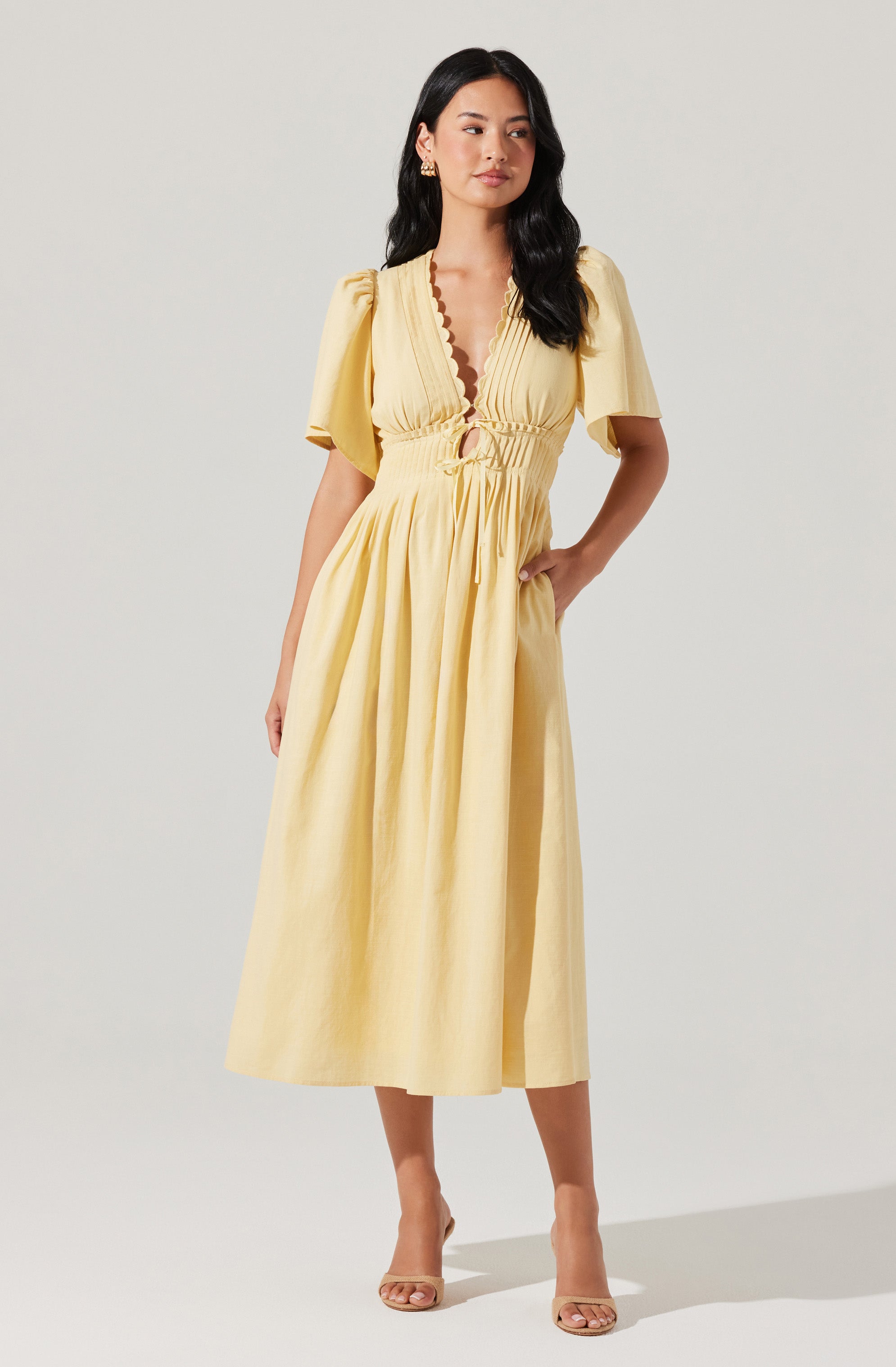 A woman is wearing a long, yellow dress made of a lightweight, breezy fabric. The dress features short puffed sleeves, a deep V-neckline with a scalloped edge, and a drawstring waist that creates a gathered effect. The skirt is full and flows down to her ankles. She is also wearing nude high-heeled sandals.