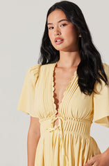 A woman is wearing a light yellow dress with short puffed sleeves. The dress features a deep V neckline with a scalloped trim, and there are vertical pleats along the bodice. The waistline is cinched with a gathered elastic band and a matching fabric tie at the front.