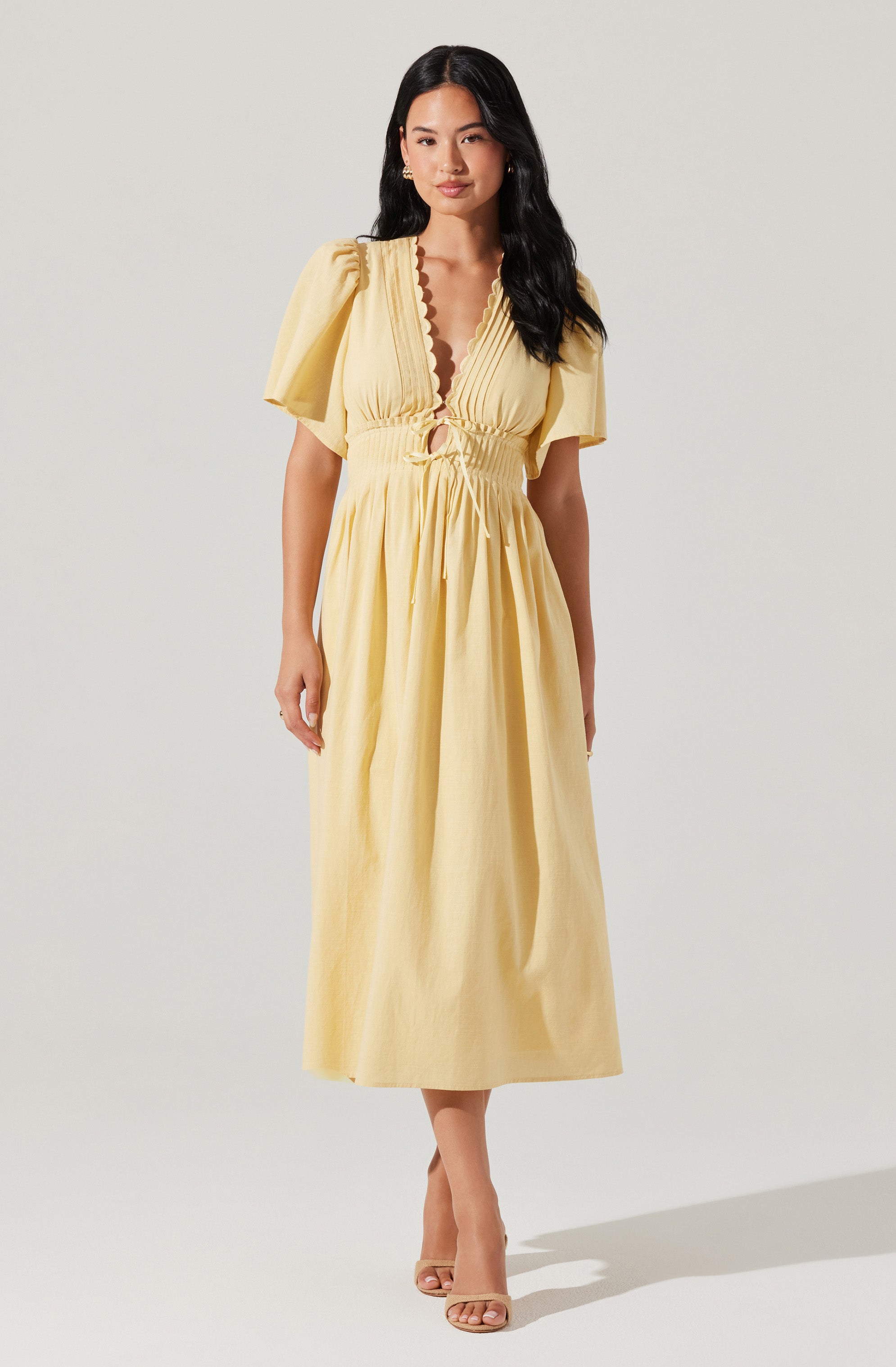 A woman is wearing a yellow maxi dress with puffed short sleeves, a deep V neckline with a scalloped edge, and a gathered, ruched waist with a tie-up detail. The dress has a flowing skirt that reaches the ankles.