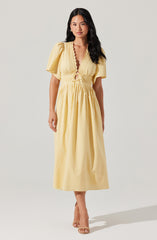 A woman is wearing a yellow maxi dress with puffed short sleeves, a deep V neckline with a scalloped edge, and a gathered, ruched waist with a tie-up detail. The dress has a flowing skirt that reaches the ankles.