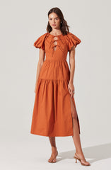 A woman is wearing a long, burnt orange dress with short, puffy sleeves. The dress features a fitted bodice with a lace-up detail at the front, including oval cutouts. The skirt is gathered at the waist, with a high slit on one side. She is also wearing nude, open-toe heels.