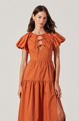 A woman is wearing a coral-colored dress with puffed short sleeves, a fitted bodice with a lace-up front featuring multiple bows, and a tiered skirt with a slit on one side.