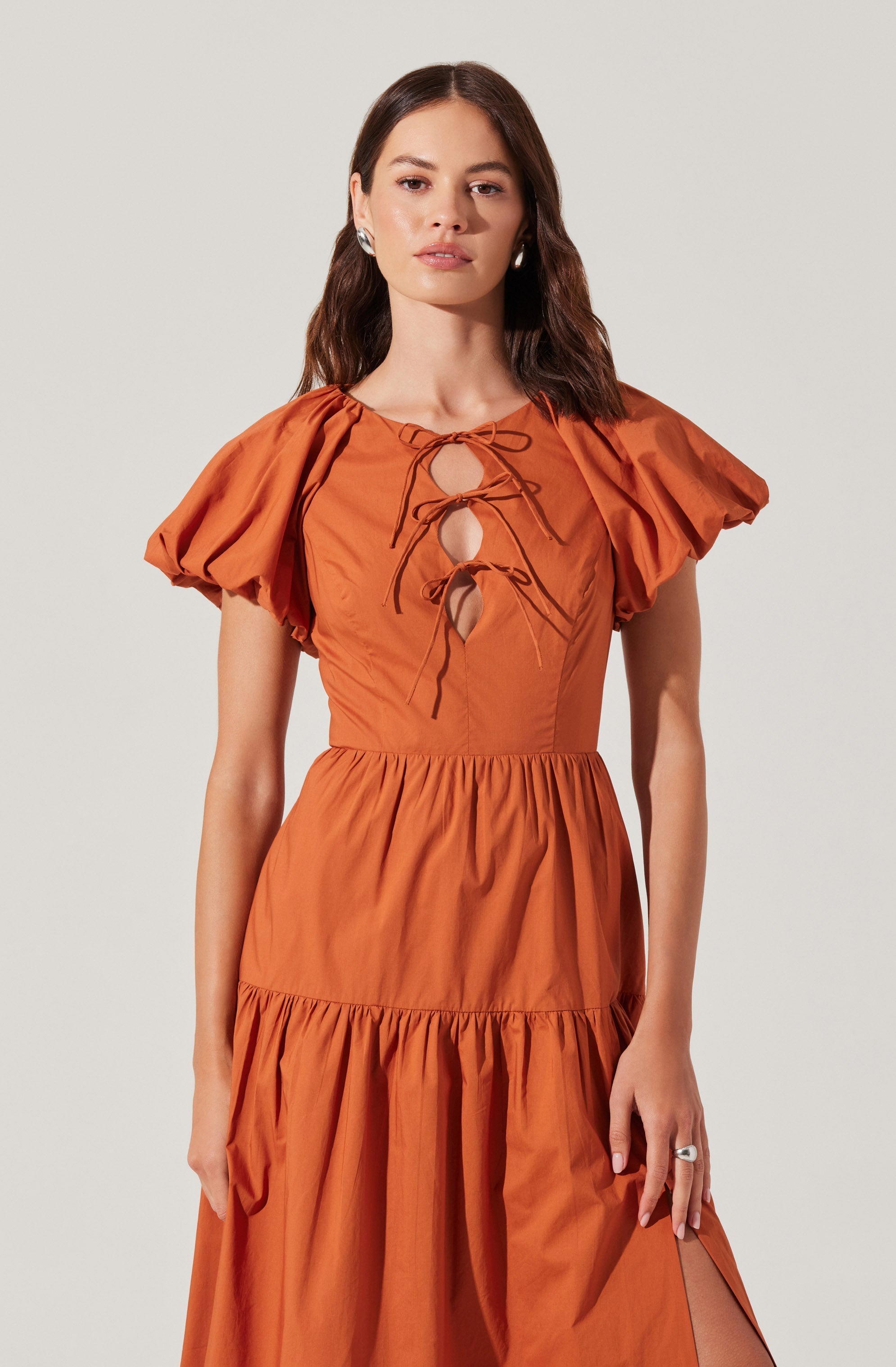 A woman is wearing a burnt orange dress with puffed short sleeves, a fitted waist, and a tiered skirt. The dress has a round neckline with a keyhole opening in the front, accented by a drawstring tie that creates a bow.