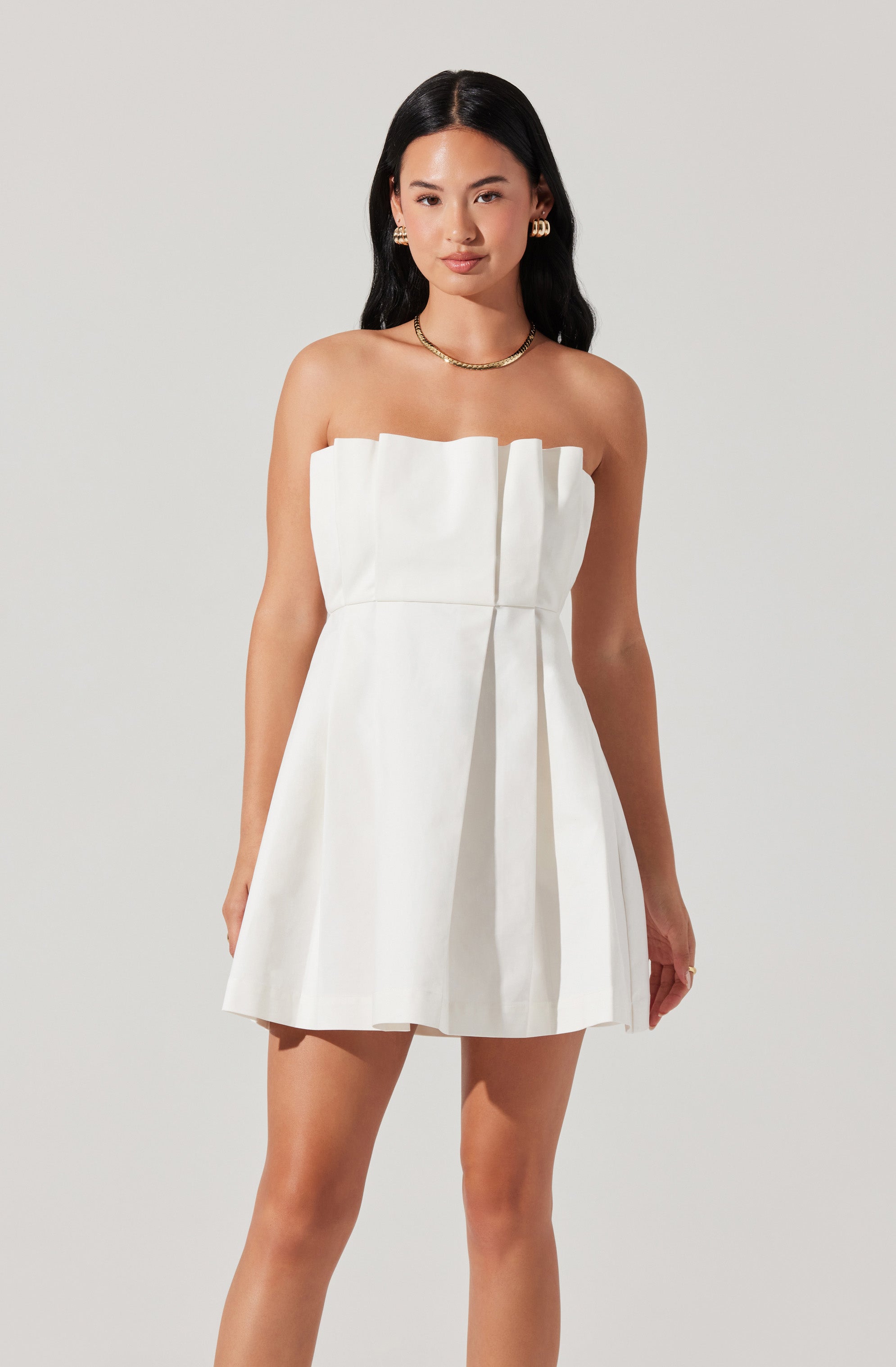 A woman is wearing a strapless white dress with a structured, pleated bodice and a slightly flared skirt. The dress features a prominent front pleat detail that runs vertically down the center. She accessorizes with gold jewelry, including a necklace, earrings, and rings.