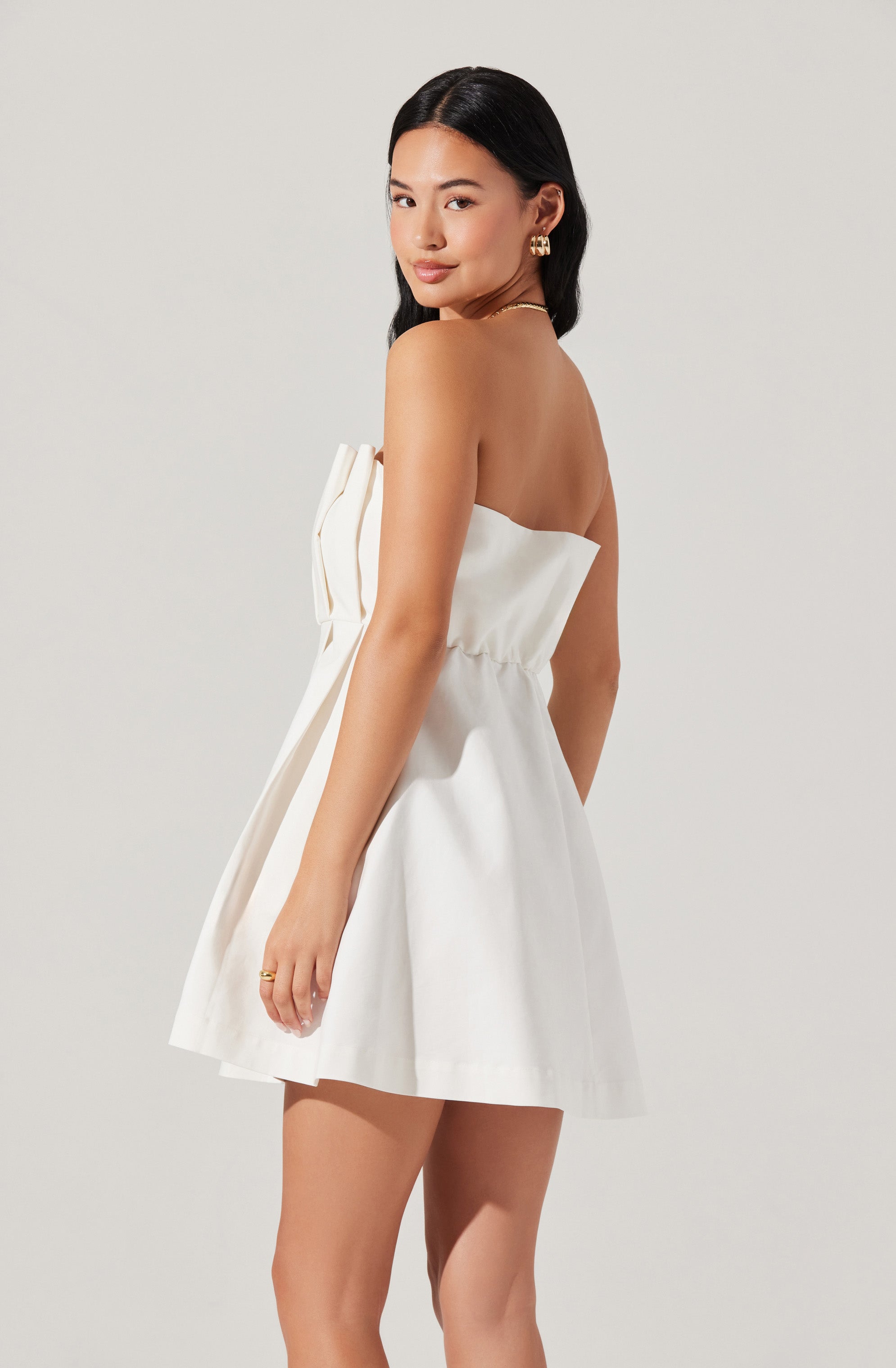 A woman is wearing a strapless white dress with a fitted waist and a short, flared skirt. The dress features a large bow or draped detail on the front. She accessorizes with gold earrings and a delicate necklace.