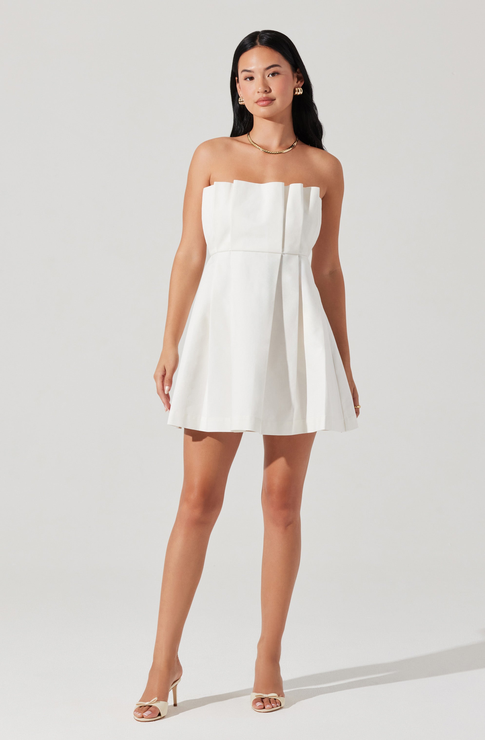 A woman is wearing a strapless white dress with a pleated skirt. The dress has a straight neckline with a ruffled detail at the top. She is accessorized with gold jewelry, including hoop earrings, a necklace, and rings. She is also wearing strappy high-heeled sandals.