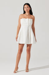 A woman is wearing a strapless white dress with a pleated skirt. The dress has a straight neckline with a ruffled detail at the top. She is accessorized with gold jewelry, including hoop earrings, a necklace, and rings. She is also wearing strappy high-heeled sandals.
