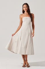 The dress is a strapless, knee-length, A-line style made from a light, ivory-colored fabric. It has a fitted bodice with horizontal seam detailing at the top and vertical seams running down the skirt, creating a slightly pleated effect. The fabric appears smooth and structured, offering a classic and elegant look. The woman is accessorized with gold jewelry, including a necklace and earrings, and is wearing black strappy high-heeled sandals.