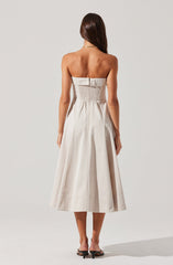 A woman is wearing a strapless, knee-length dress in a light, neutral color. The dress features a fitted bodice with ruching and gathers at the waist, creating a draped effect. The skirt is full with pleats that give it volume and a subtle sheen. The woman is also wearing open-toe heeled sandals and a delicate necklace.