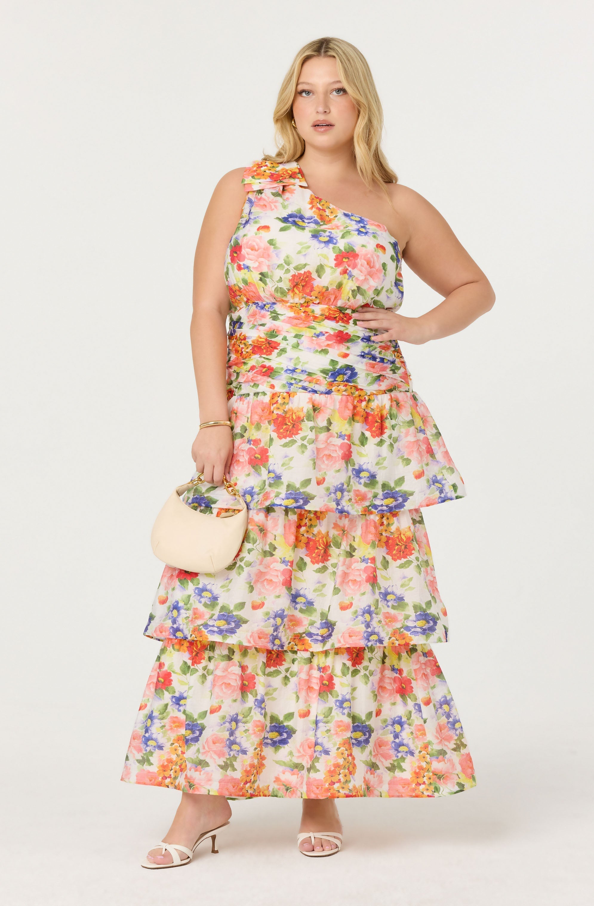 A woman is wearing a sleeveless, tiered maxi dress with a colorful floral pattern featuring pink, orange, purple, and green flowers on a light background.