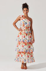 A woman is wearing a sleeveless, asymmetrical floral dress with ruffle details. The dress features a mix of vibrant colors including pink, orange, purple, and green, with a tiered skirt that has multiple layers, each with ruffled edges. She is also wearing a gold bracelet and open-toed sandals.