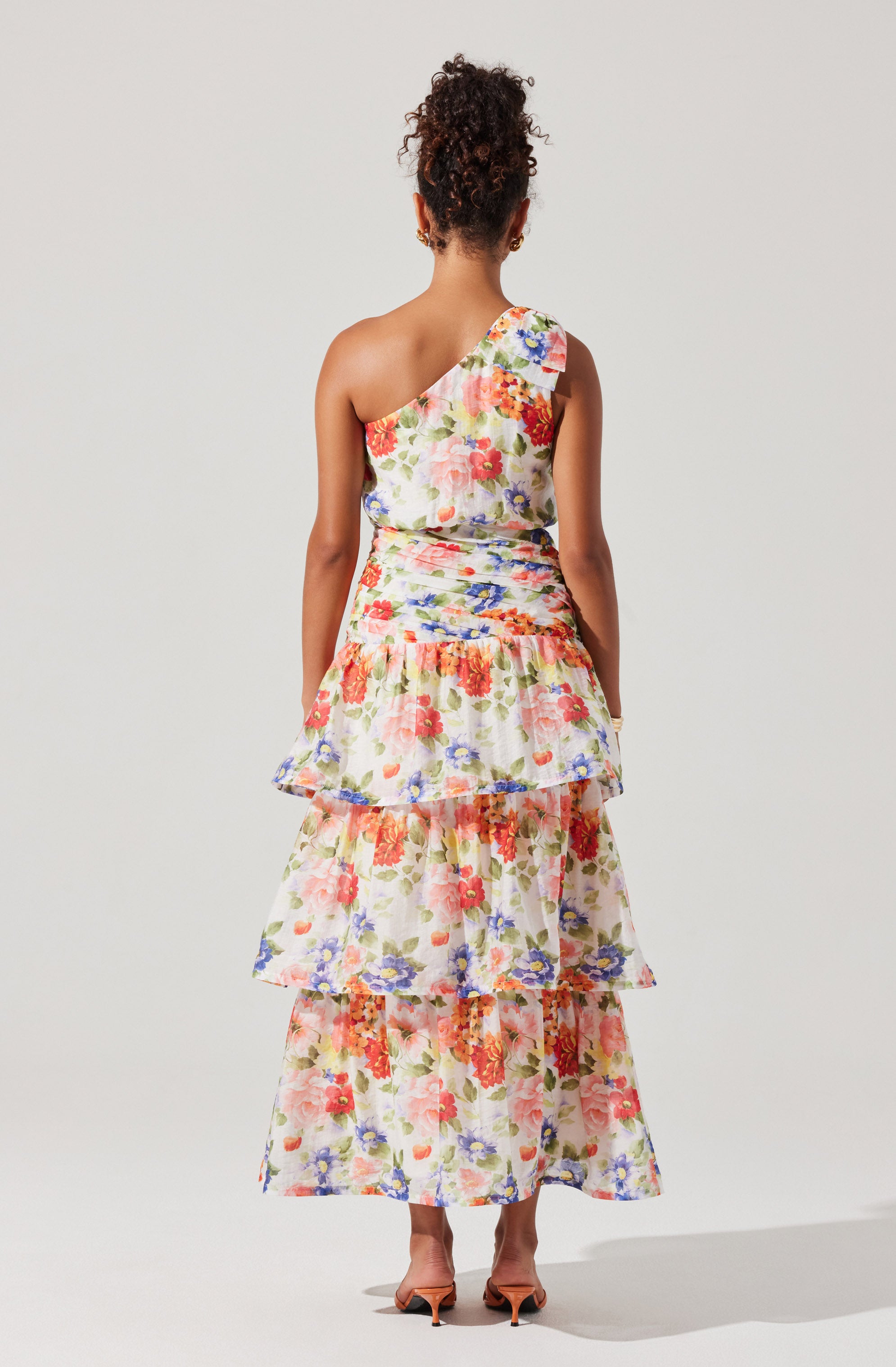 A woman is wearing a sleeveless, one-shoulder dress with a floral print in shades of pink, red, purple, and green on a light background. The dress features multiple tiered ruffles that create a layered effect, and the fabric appears textured with a slight ruching detail around the waist. The woman is also wearing open-toe high heels.