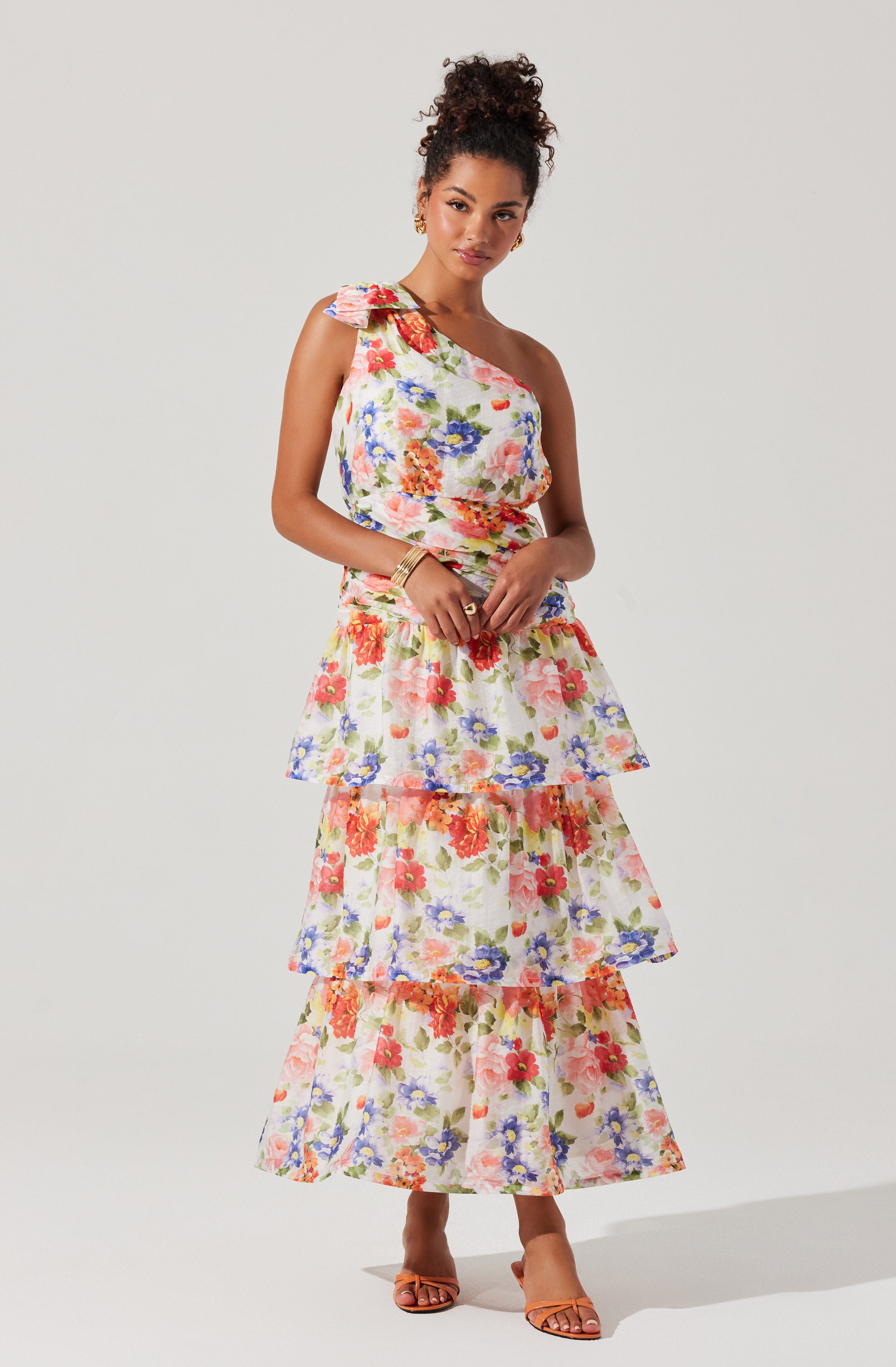 A woman is wearing a sleeveless, one-shoulder dress with ruffled details on the shoulder. The dress features a floral pattern with large, colorful flowers in shades of pink, orange, purple, and blue on a light background. The dress has a tiered, ruffled skirt that extends to her ankles. She is also wearing open-toe heels and accessories including bracelets.