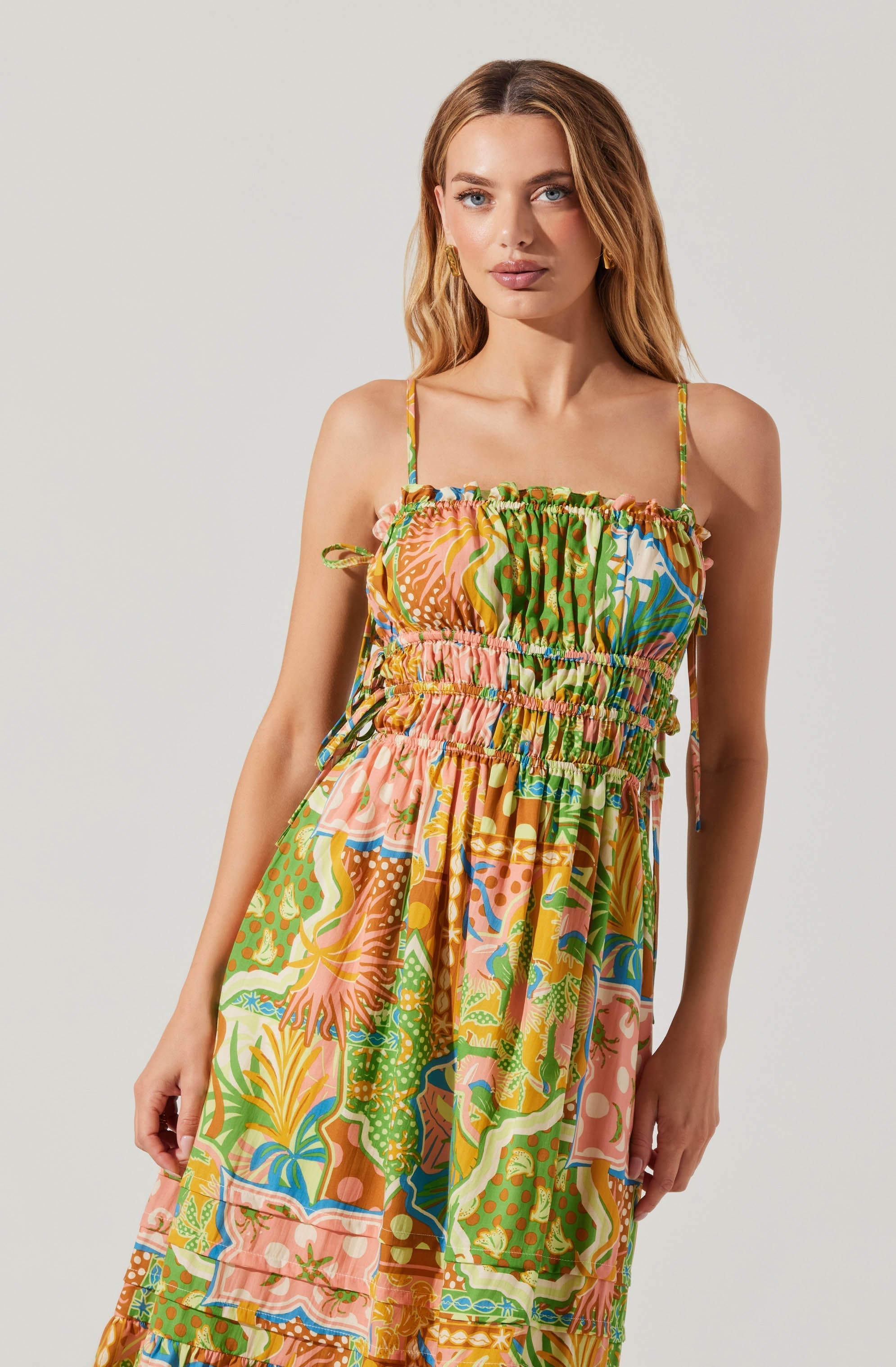 A woman wearing a colorful, patterned dress with thin spaghetti straps. The dress features an elastic, smocked bodice and a flowing skirt with vibrant, abstract and floral designs in shades of green, pink, yellow, blue, and orange.