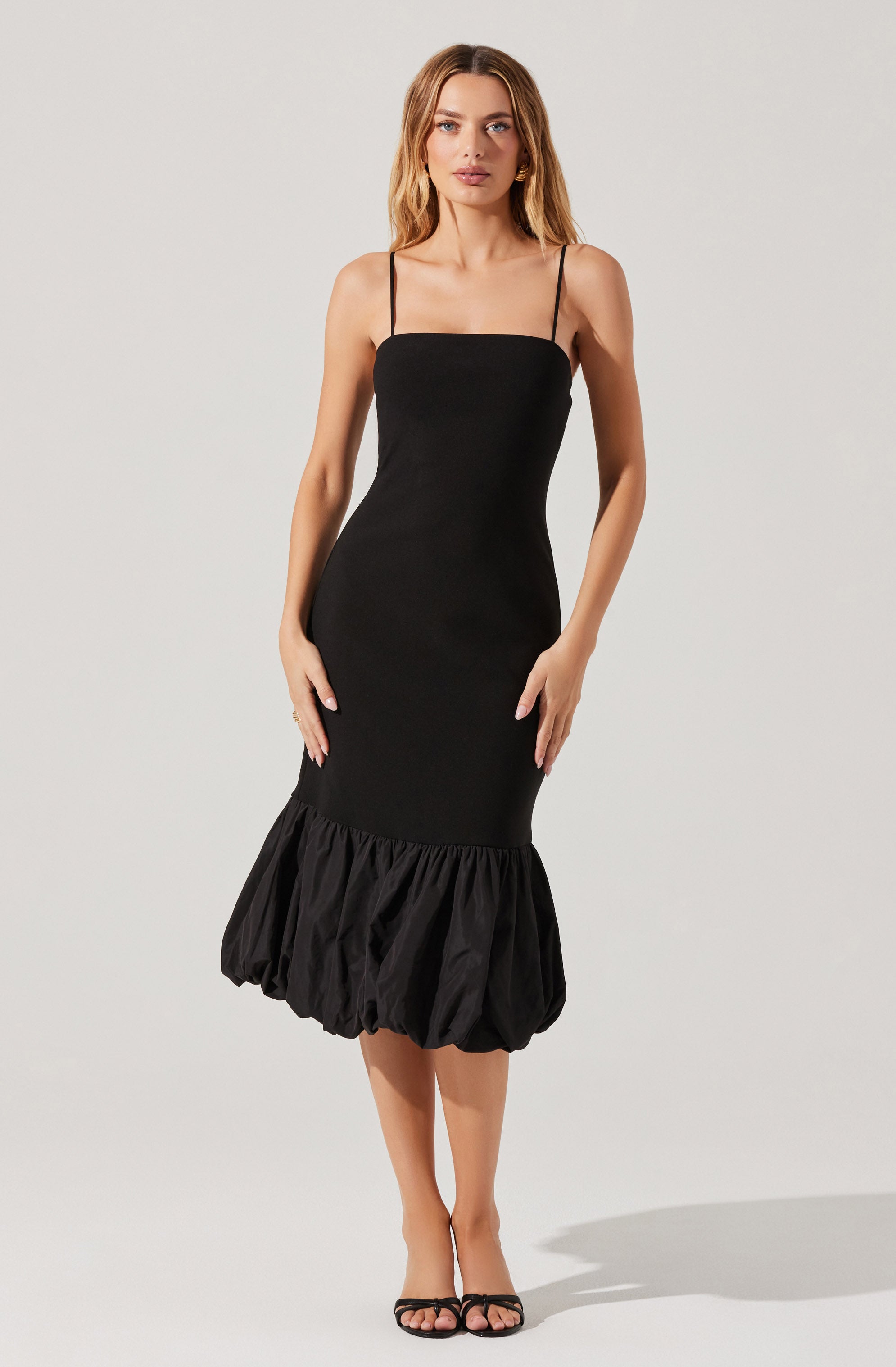 A woman is wearing a black sleeveless dress with thin spaghetti straps. The dress has a fitted bodice and a gathered, voluminous bubble hem at the bottom. She is also wearing black open-toe high-heeled shoes.