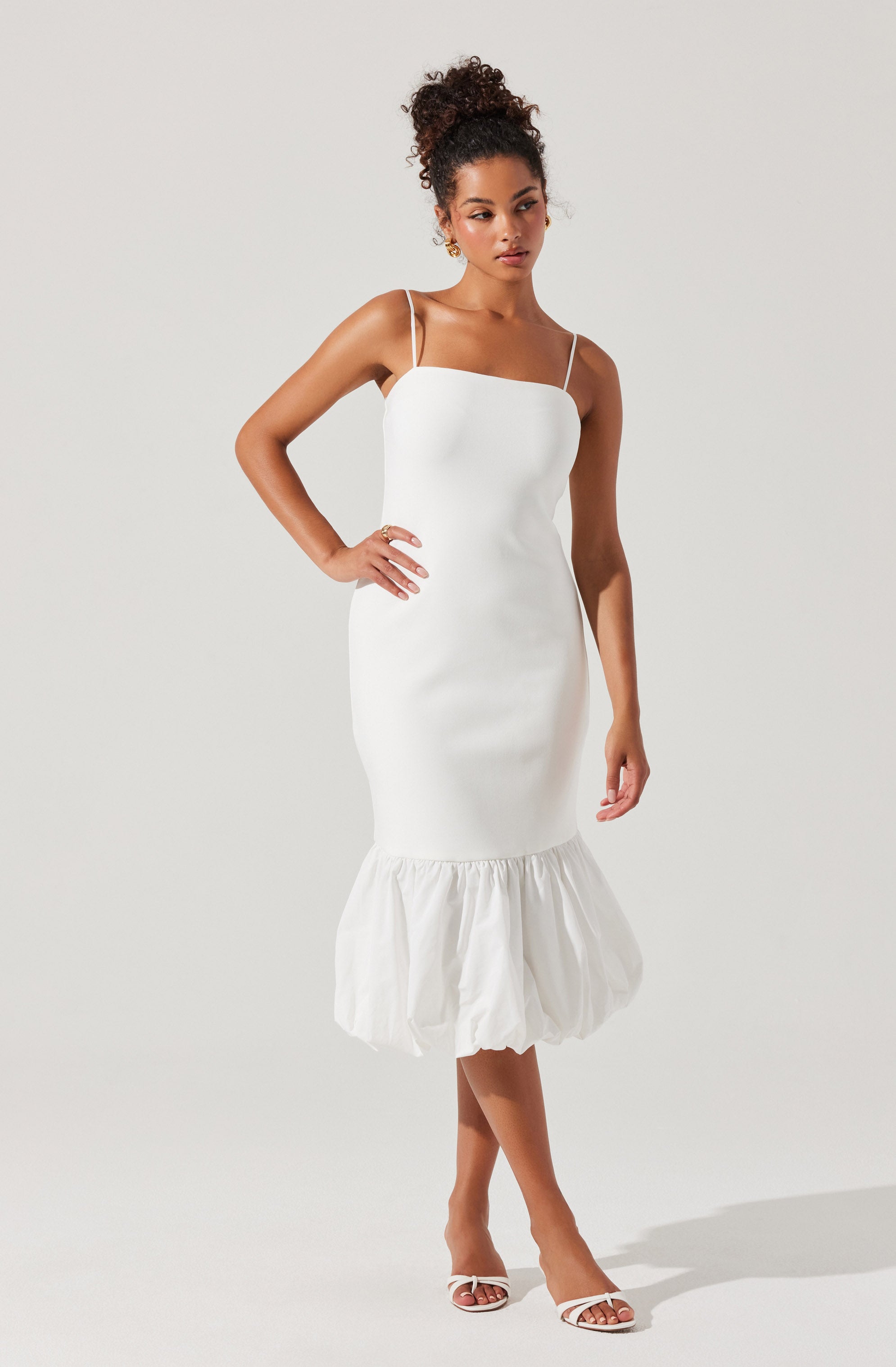 A woman is wearing a white dress with thin spaghetti straps. The dress is fitted through the torso and hips, then features a ruffled, gathered hem that falls below the knees. She is also wearing white heeled sandals.