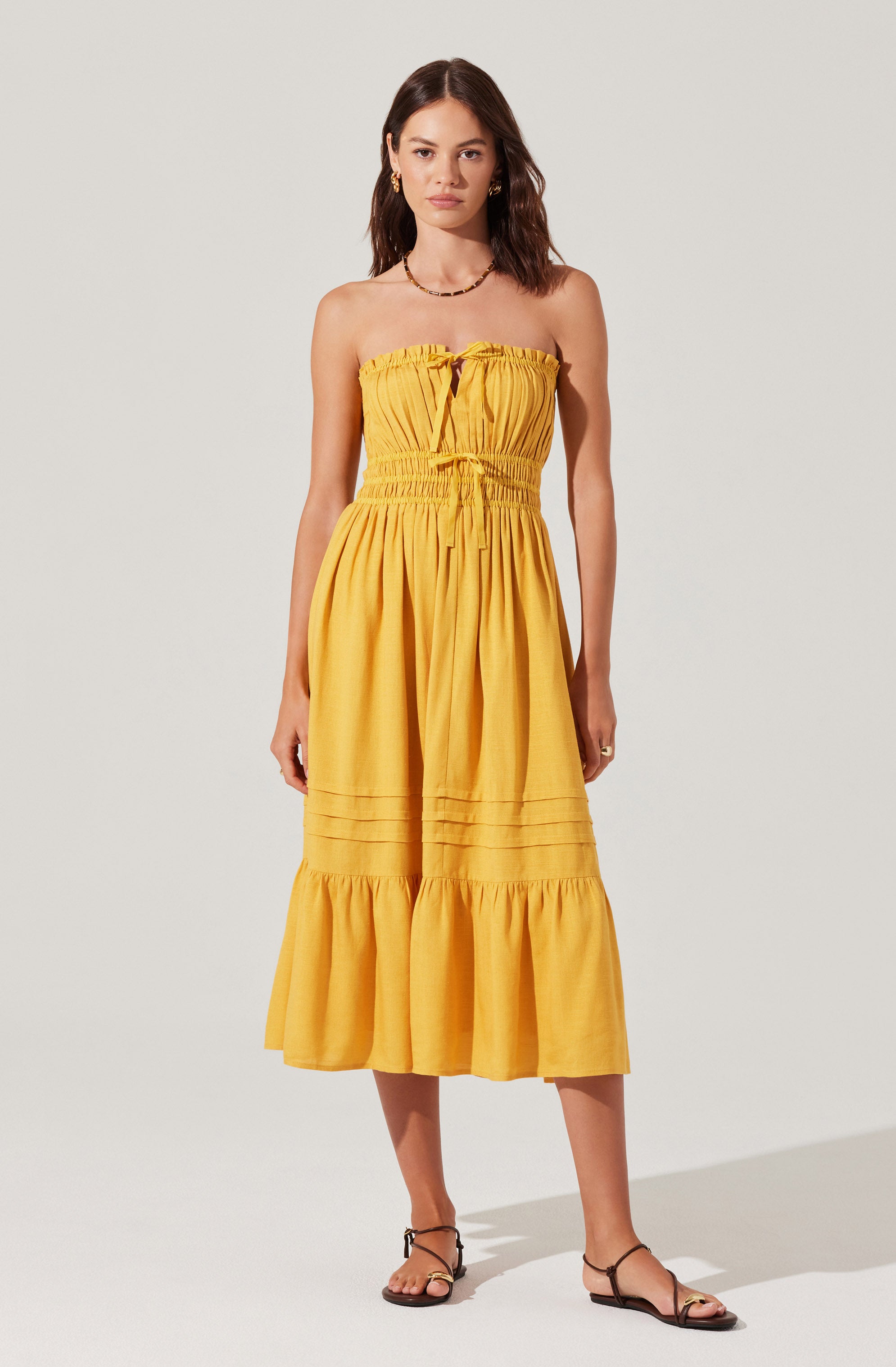 A young woman is wearing a strapless, yellow midi dress with a smocked bodice that has a tie detail at the neckline. The dress features a gathered waist, vertical pleats down the skirt, and a ruffled hem that falls below the knees. She is accessorized with a necklace, earrings, and a ring, and is wearing black strappy sandals.