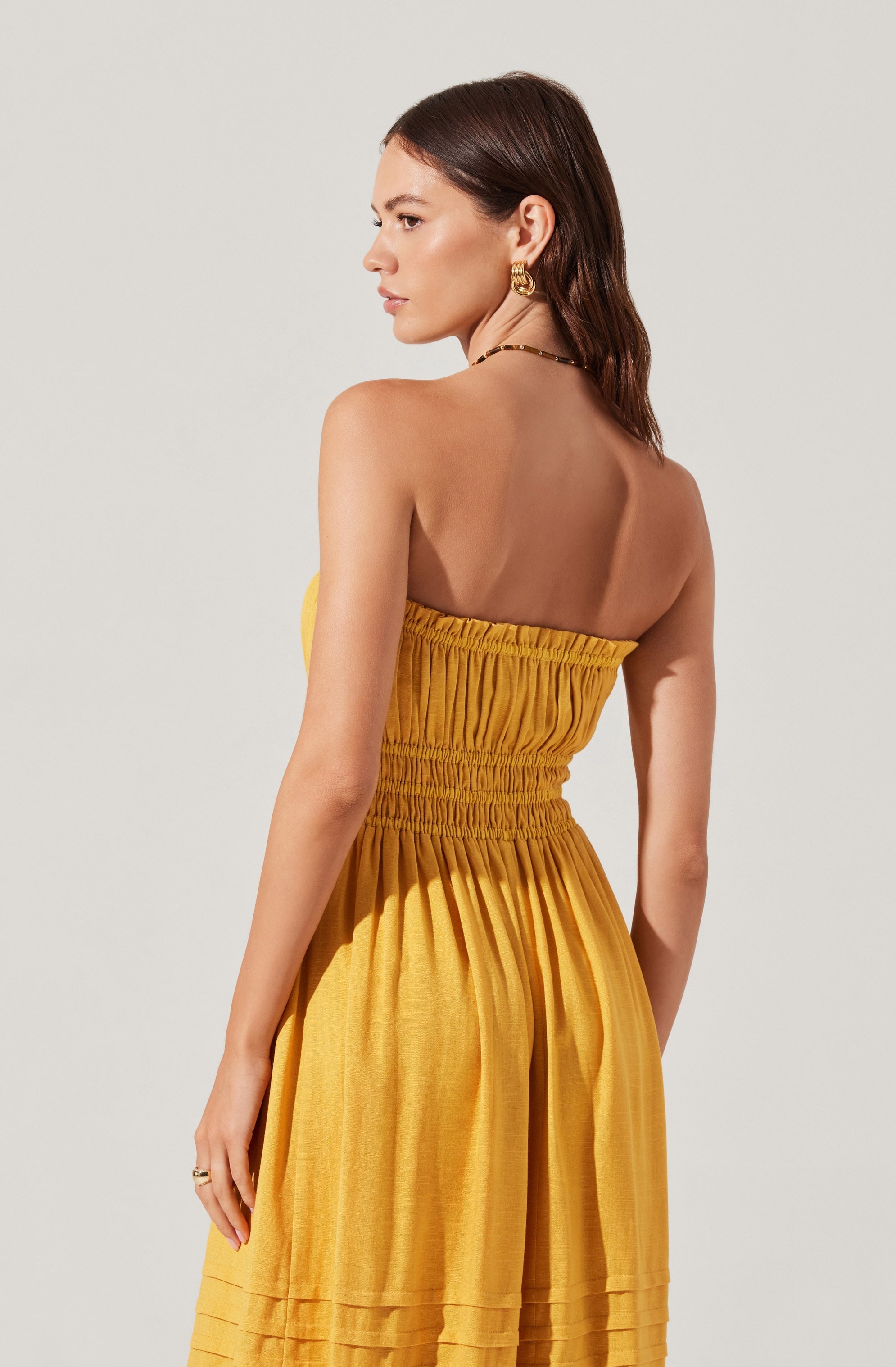 A woman is wearing a strapless, bright yellow dress with a smocked bodice and elastic waistband. The dress is flowy with gathered fabric at the waist and pleated details near the hem. She accessorizes with large gold hoop earrings, a bracelet, and a necklace. Her hair is styled in soft waves, and she has a neutral makeup look.
