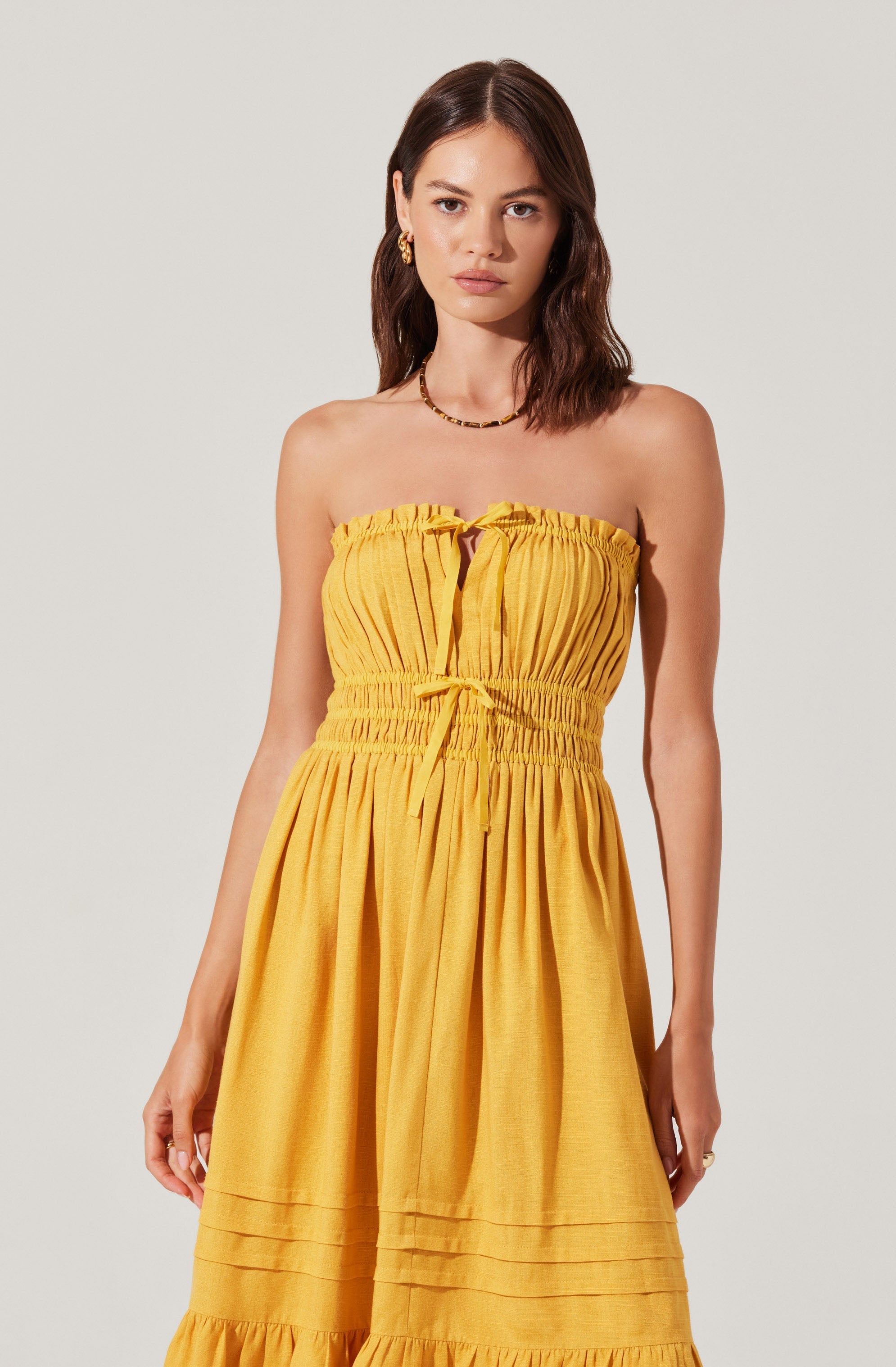 A woman is wearing a strapless, bright yellow dress with gathered fabric at the bust and waist. The dress features vertical pintuck details on the bodice and horizontal seam details with pleats near the hemline. The fabric appears lightweight and textured, giving the dress a summery, casual look. She accessorizes with small hoop earrings and a delicate necklace.
