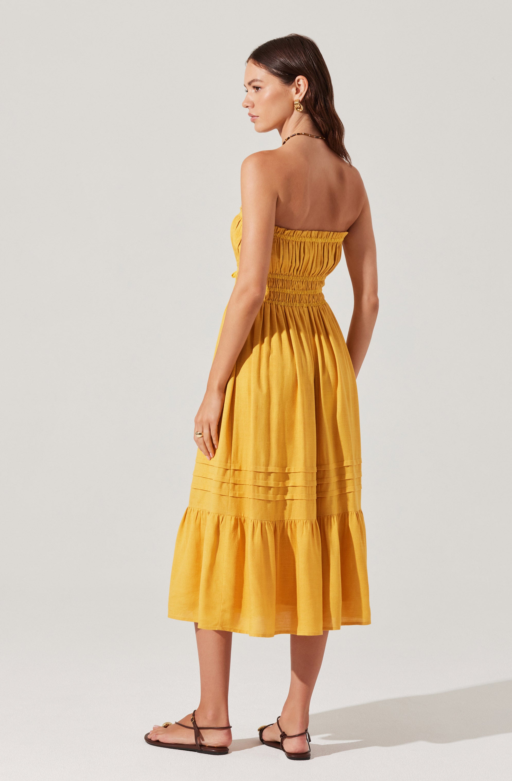 A woman is wearing a strapless, yellow, tiered midi dress with ruched detailing at the bodice and gathered fabric at the hem. She has on simple, flat, brown sandals, gold hoop earrings, and a delicate necklace.