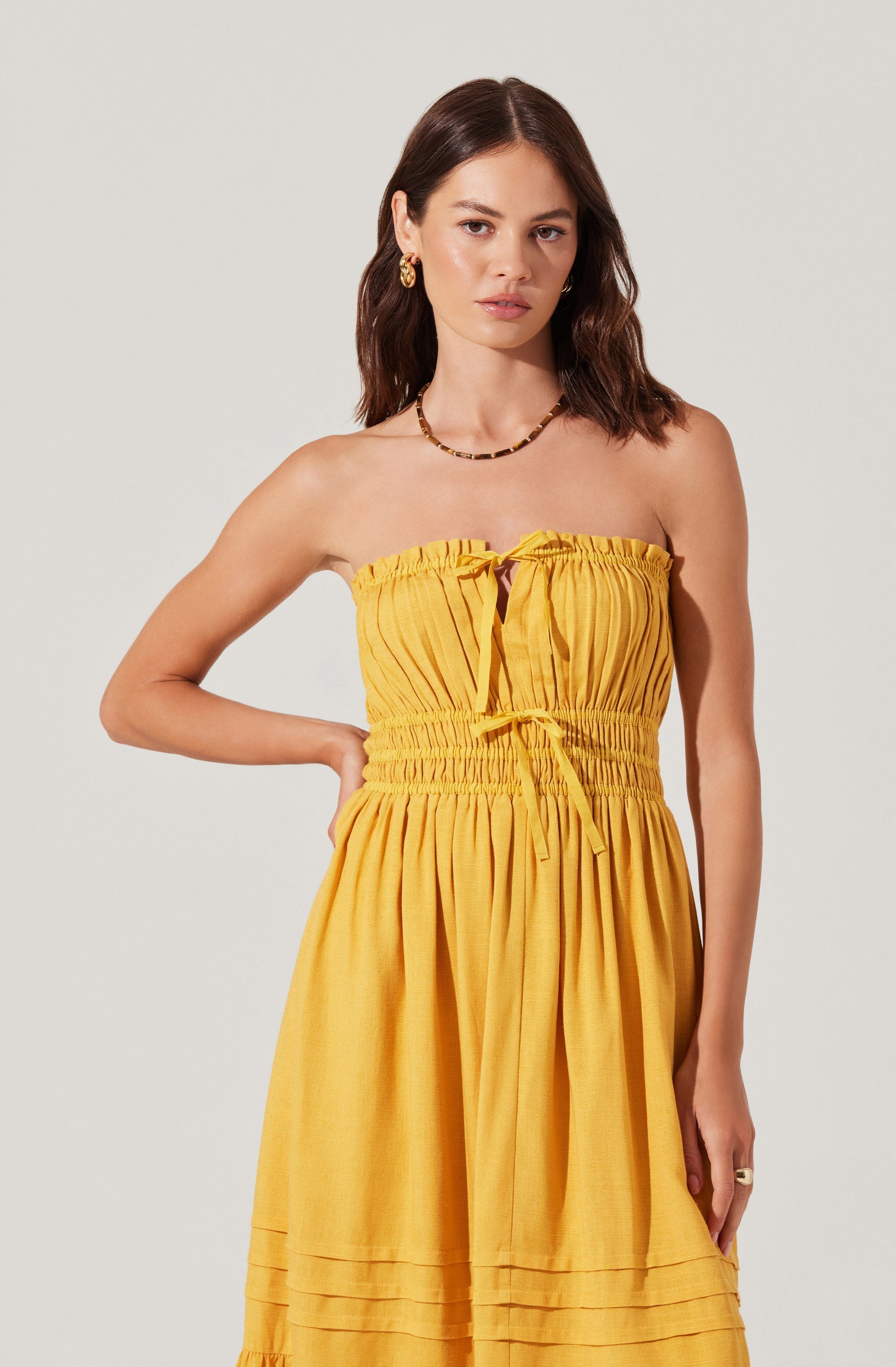 A young woman is wearing a strapless, bright yellow, gathered dress with a ruched bodice that has a drawstring tie at the center. The dress features a high waist with elastic gathering and a flowing skirt with horizontal pleats near the hem. She accessorized with gold hoop earrings, a beaded necklace, and a ring on her right hand.