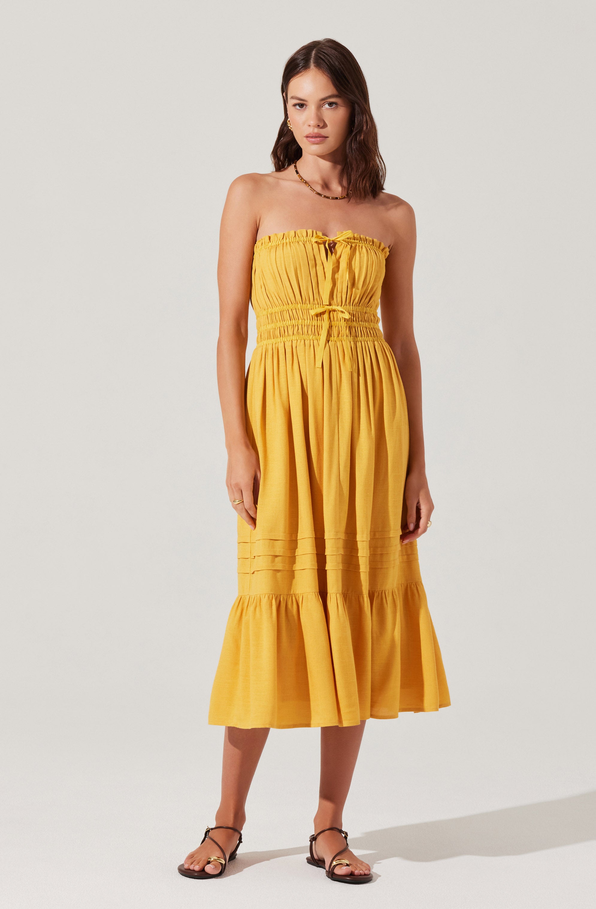 The woman is wearing a strapless yellow maxi dress with a gathered bodice that has a tie detail at the center. The dress features a cinched, elasticized waist and a tiered skirt with horizontal pleats near the bottom. She is also wearing black strappy flat sandals.
