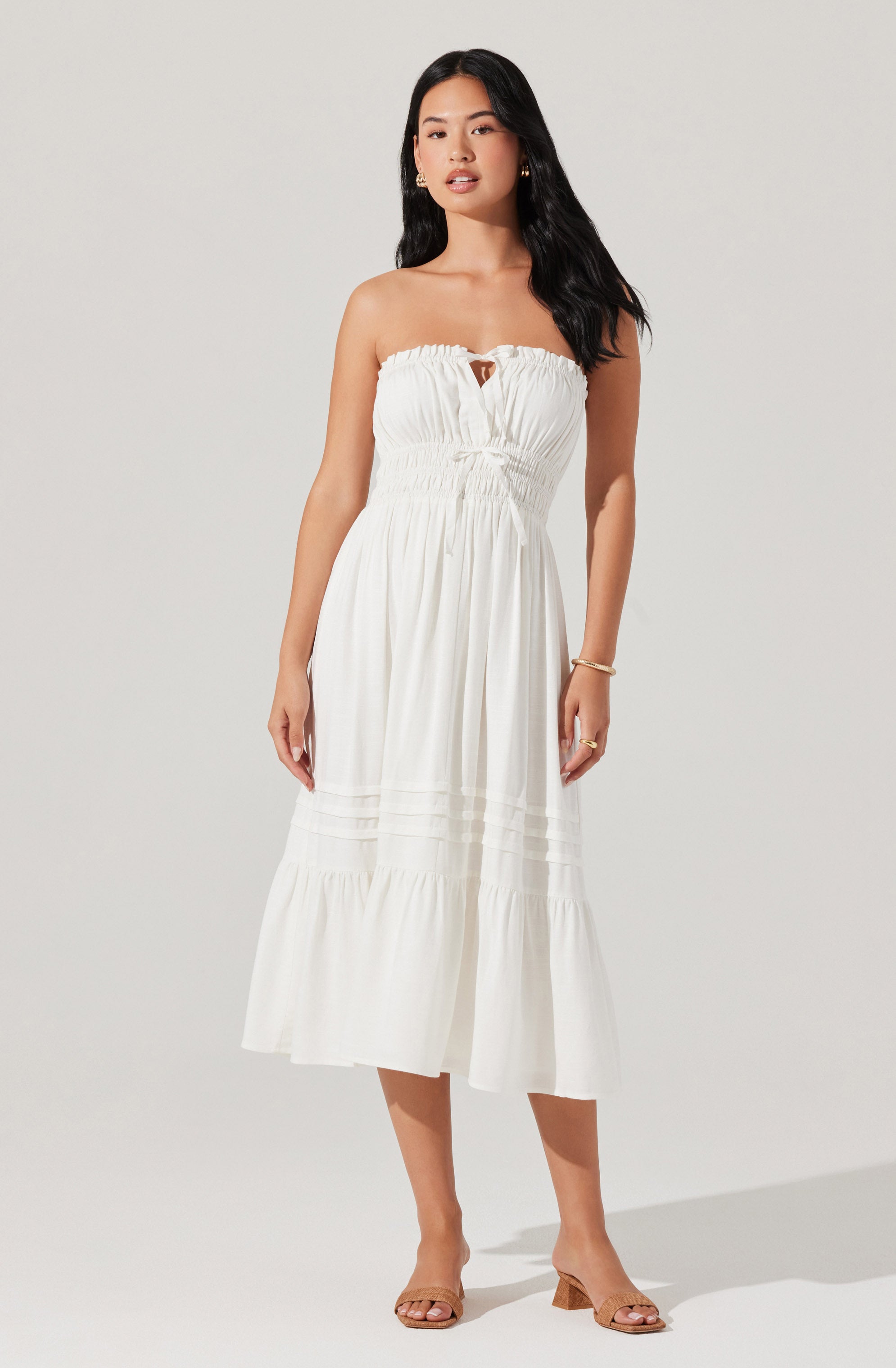 A woman is wearing a strapless white maxi dress with a ruched bodice and a small keyhole opening at the center of the neckline. The dress has a fitted waist and a flowy skirt with horizontal seam details. She is also wearing open-toed, block-heeled sandals in a neutral tone.