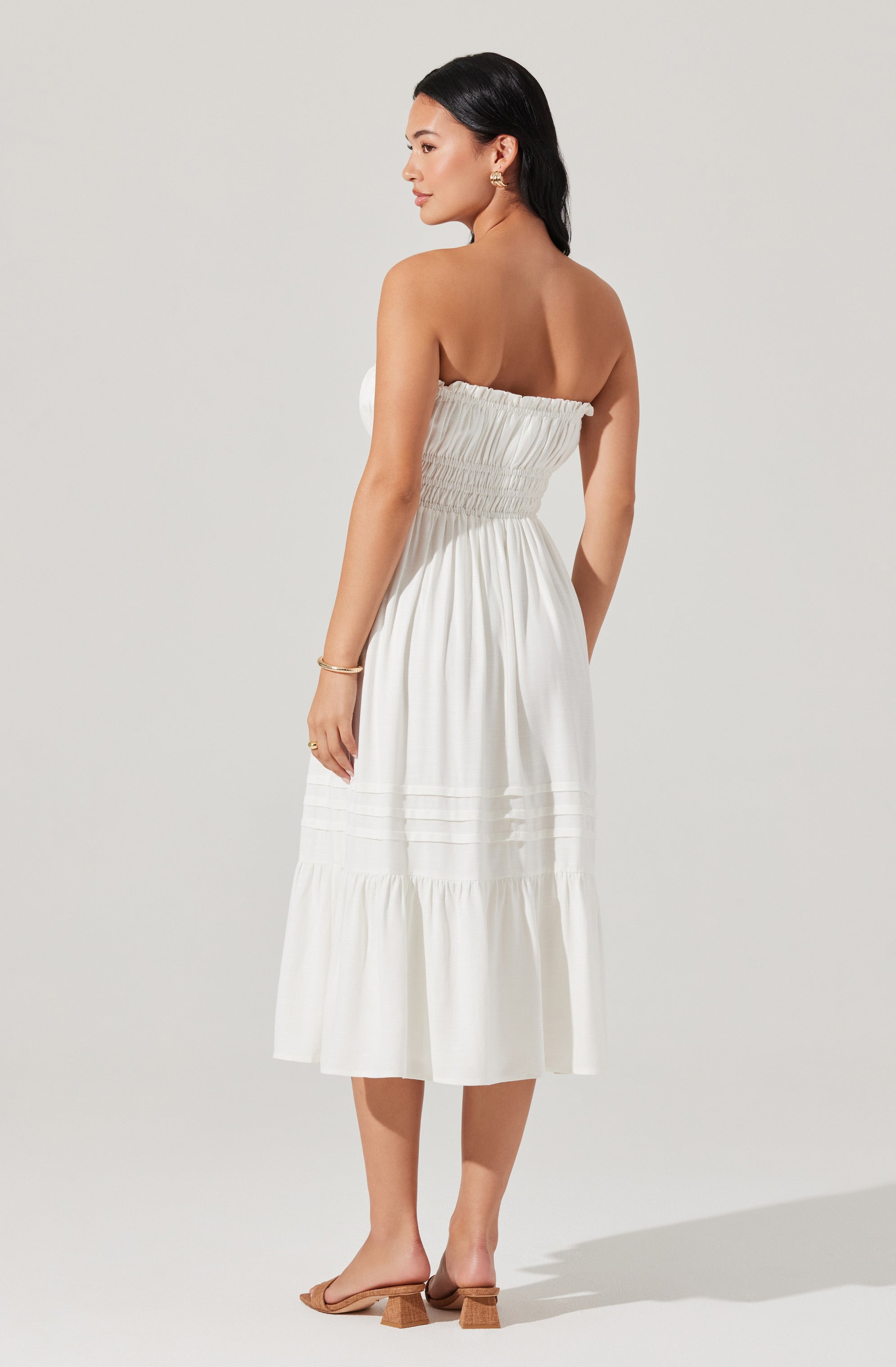 A woman is wearing a white, strapless, midi-length dress with a gathered, smocked bodice and tiered, flowing skirt. She has paired the dress with beige, open-toe, block-heeled sandals and simple jewelry, including earrings, a bangle, and a ring.