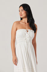 A woman is wearing a strapless white dress with a gathered, elasticized bodice, featuring a small keyhole opening in the center with a tie detail. The fabric of the dress is lightweight and flowing, with subtle horizontal pleats near the hem. She has long, wavy black hair and is standing against a plain, light-colored background.
