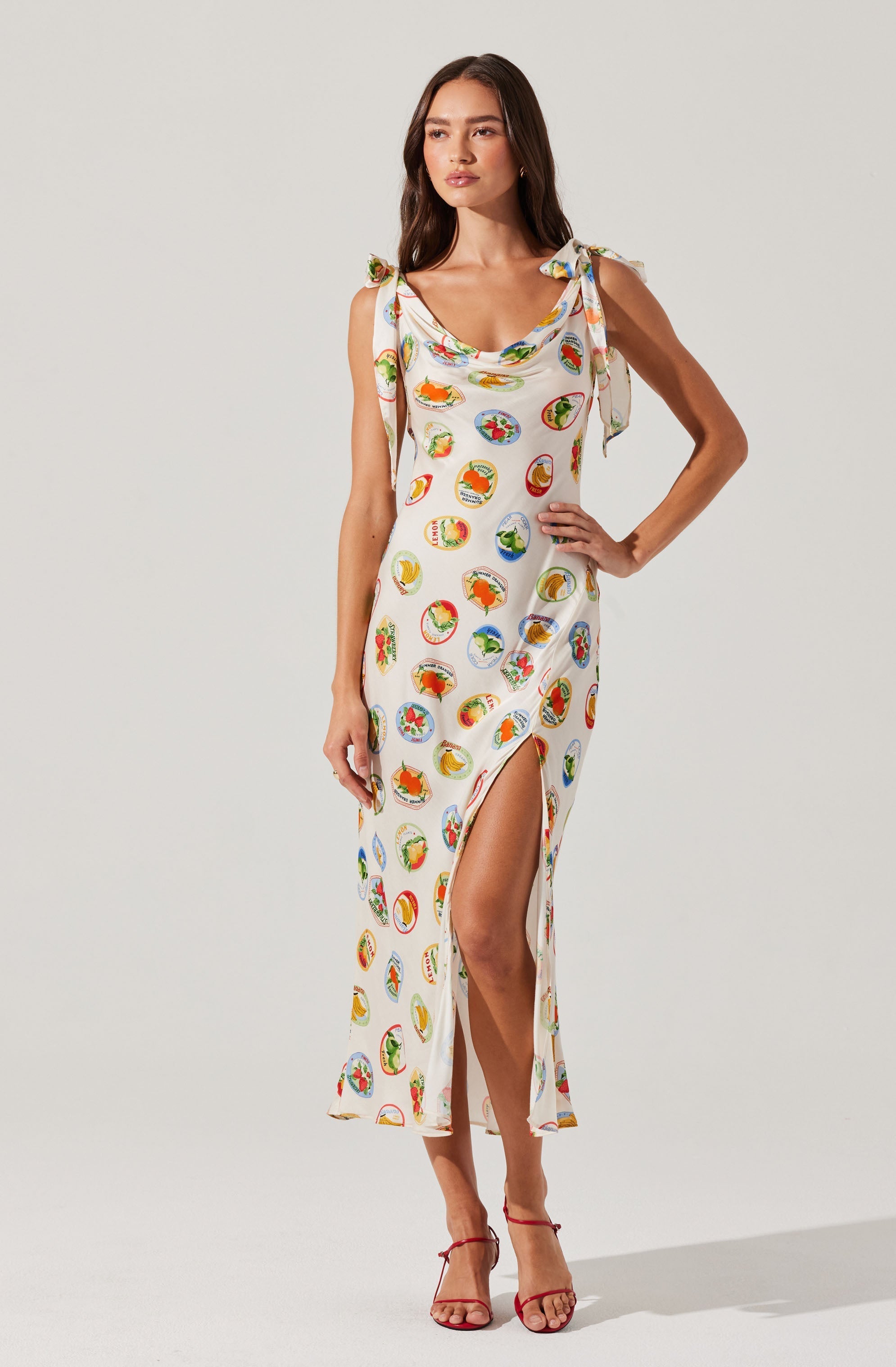 A woman wearing a sleeveless, long white dress adorned with colorful, circular fruit prints. The dress features shoulder ties and a high slit on the left side. She is also wearing red high-heeled sandals.