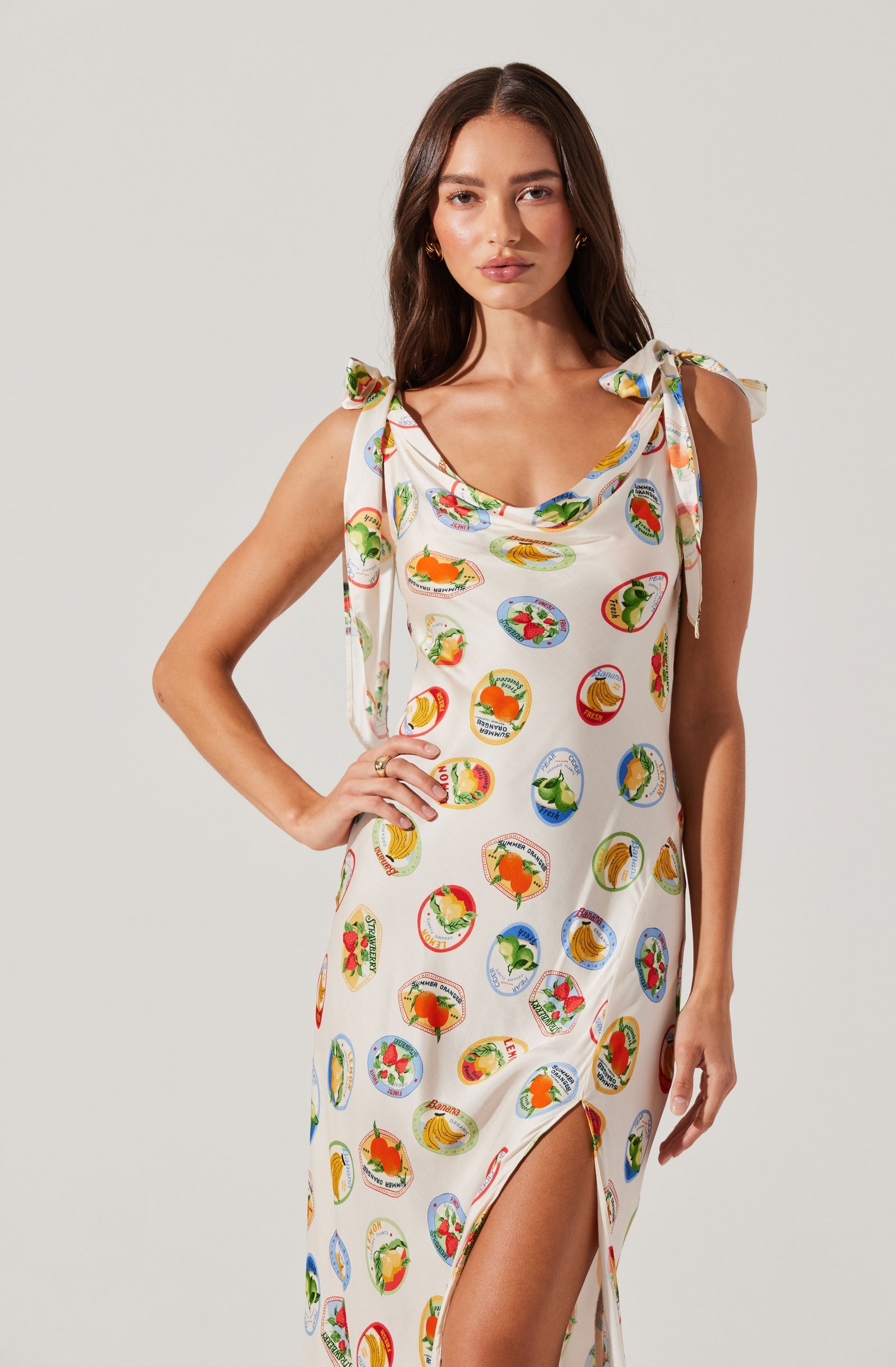 A woman is wearing a sleeveless, white dress with shoulder ties. The dress features a colorful pattern of various cartoon-style fruit and vegetable illustrations inside circular frames. The dress has a slit on the side, revealing part of her leg.