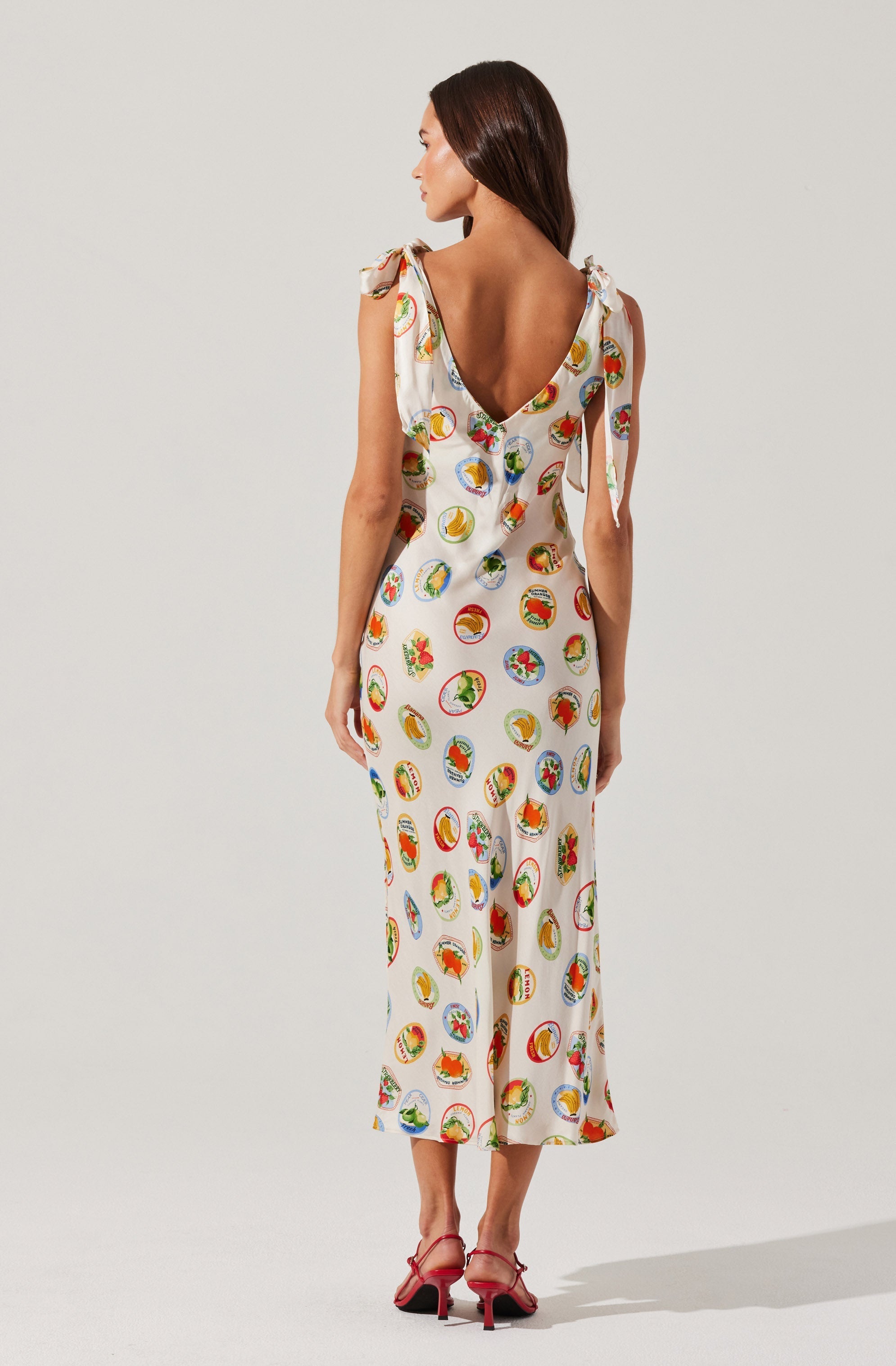 A woman is wearing a sleeveless, wide-leg jumpsuit with shoulder straps tied into bows. The fabric features a playful pattern of colorful circular illustrations, each containing various designs and images. The back of the jumpsuit has a deep V cut. She is also wearing strappy red heels.