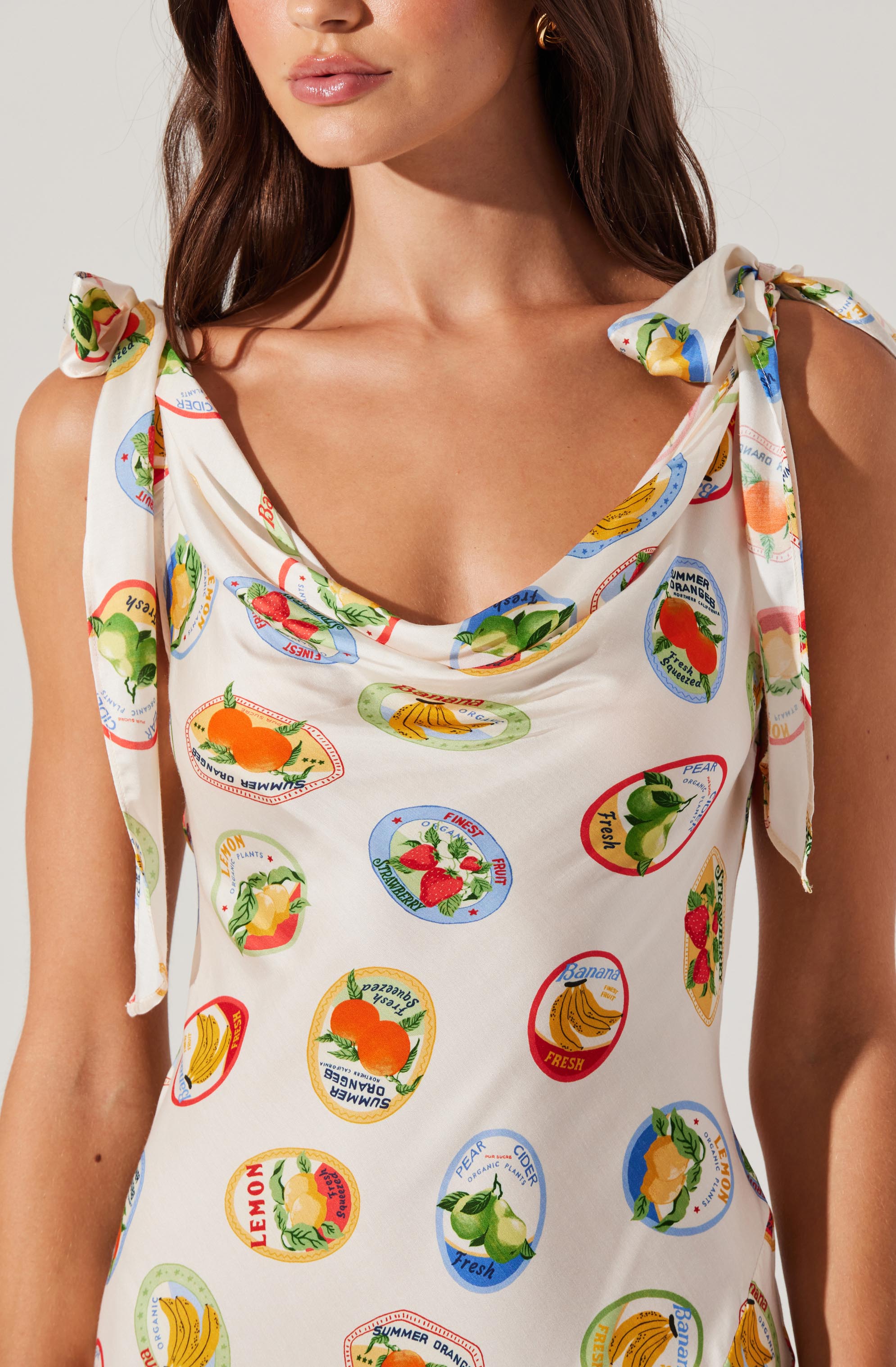 A woman is wearing a sleeveless dress with tie shoulder straps. The dress has a light-colored background and is decorated with colorful fruit-themed circular patterns, including images of bananas, strawberries, lemons, oranges, and peaches, along with words like "fresh," "summer oranges," and "fruit."