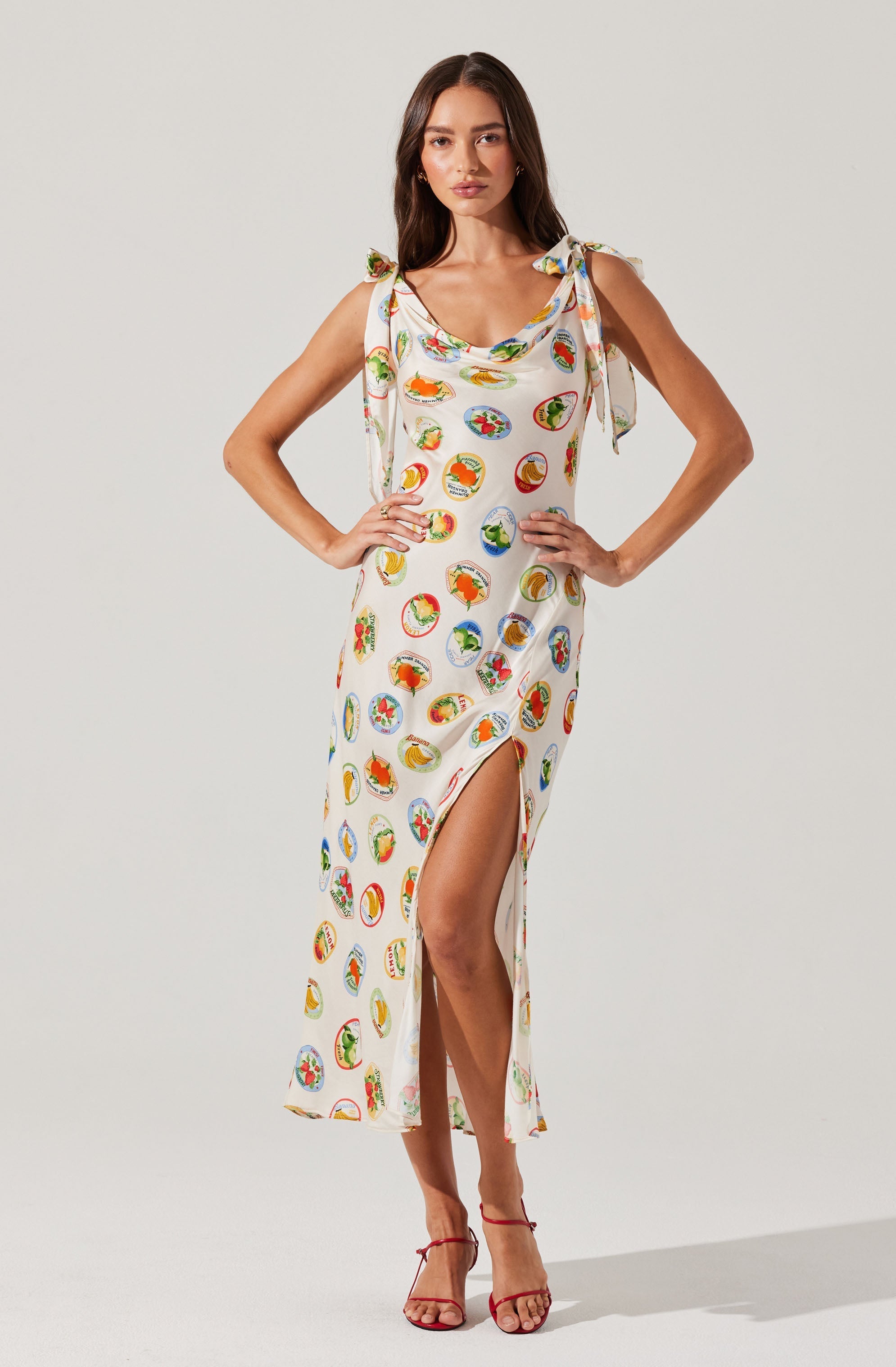 A woman is wearing a sleeveless, ankle-length dress with shoulder ties. The dress features a colorful pattern of various round, sliced fruit images printed all over. It has a relaxed fit with a side slit that reaches up to her thigh, and a scoop neckline. She pairs the dress with red strappy high-heeled sandals.