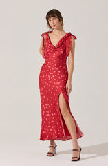 A woman is wearing a red sleeveless dress with a polka dot pattern. The dress has shoulder ties, a deep cowl neckline, and a thigh-high slit on the left side. She is also wearing black high-heeled sandals with ankle straps.